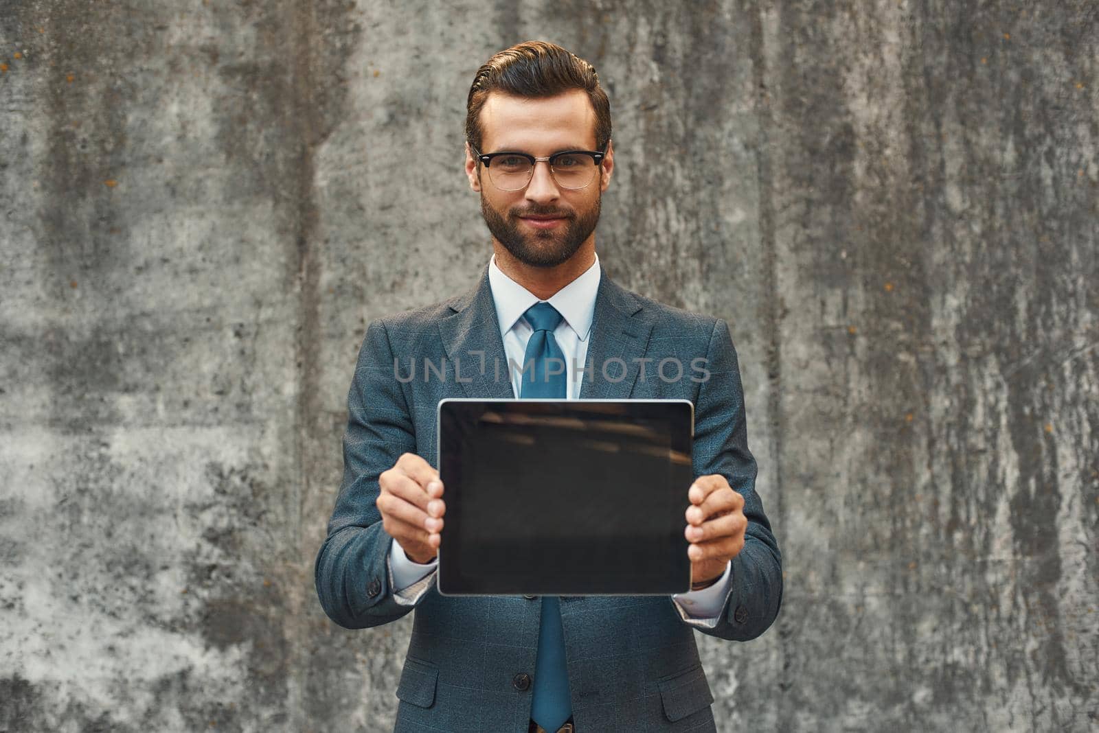 Modern businessman. Portrait of handsome bearded businessman in eyeglasses showing digital tablet at camera while standing against grey stone wall outdoors by friendsstock