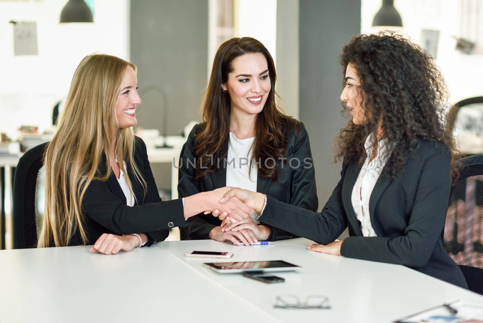 Three businesswomen shaking hands in a modern office with white furniture. Teamwork concept. Caucasian, blonde, and muslim girls wearing suit. Multi-ethnic group of women