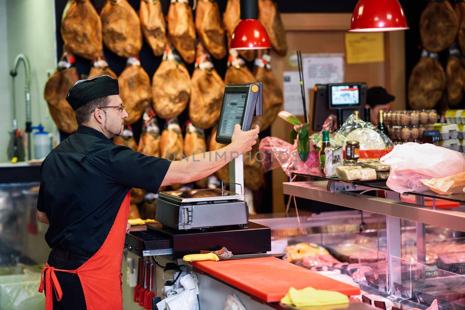 Butcher in a butcher's shop weighing the meat and charging with ham at the background