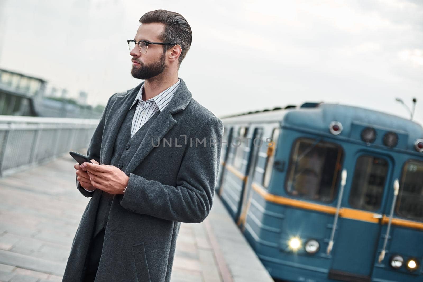 Business trip. Young businessman standing near railway holding smartphone looking aside pensive