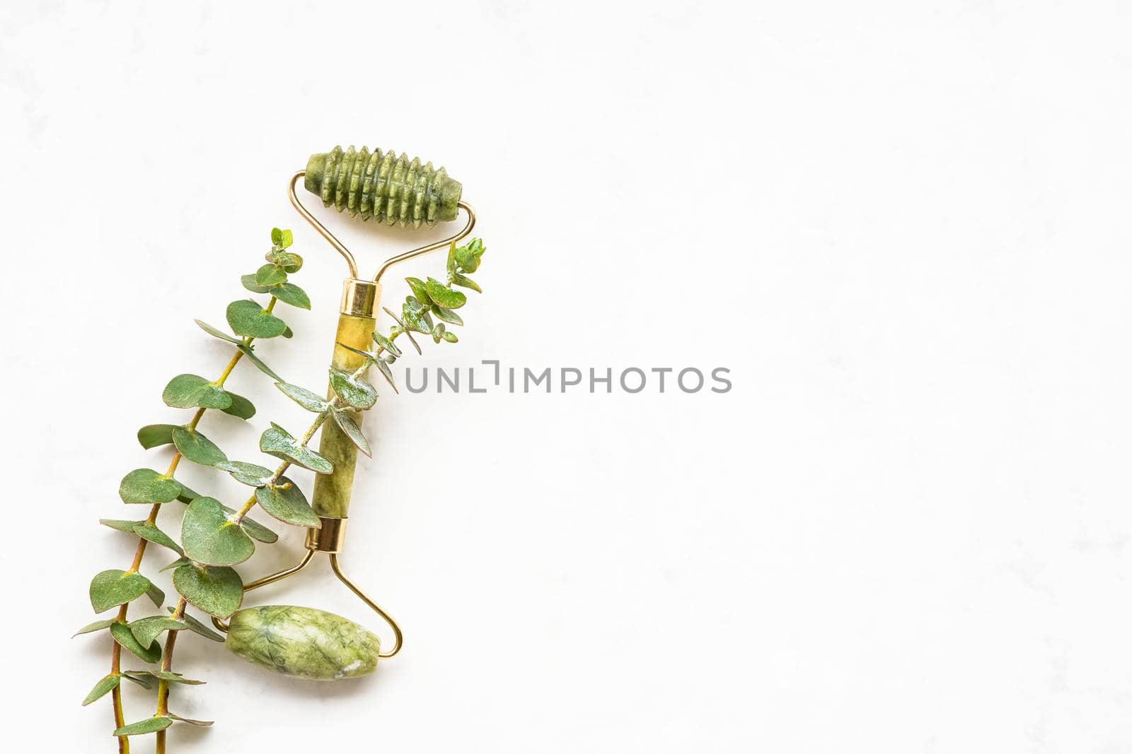 Green face roller from natural jade stone and eucalyptus branches on a white background. SPA concept. Top view, copy space for text