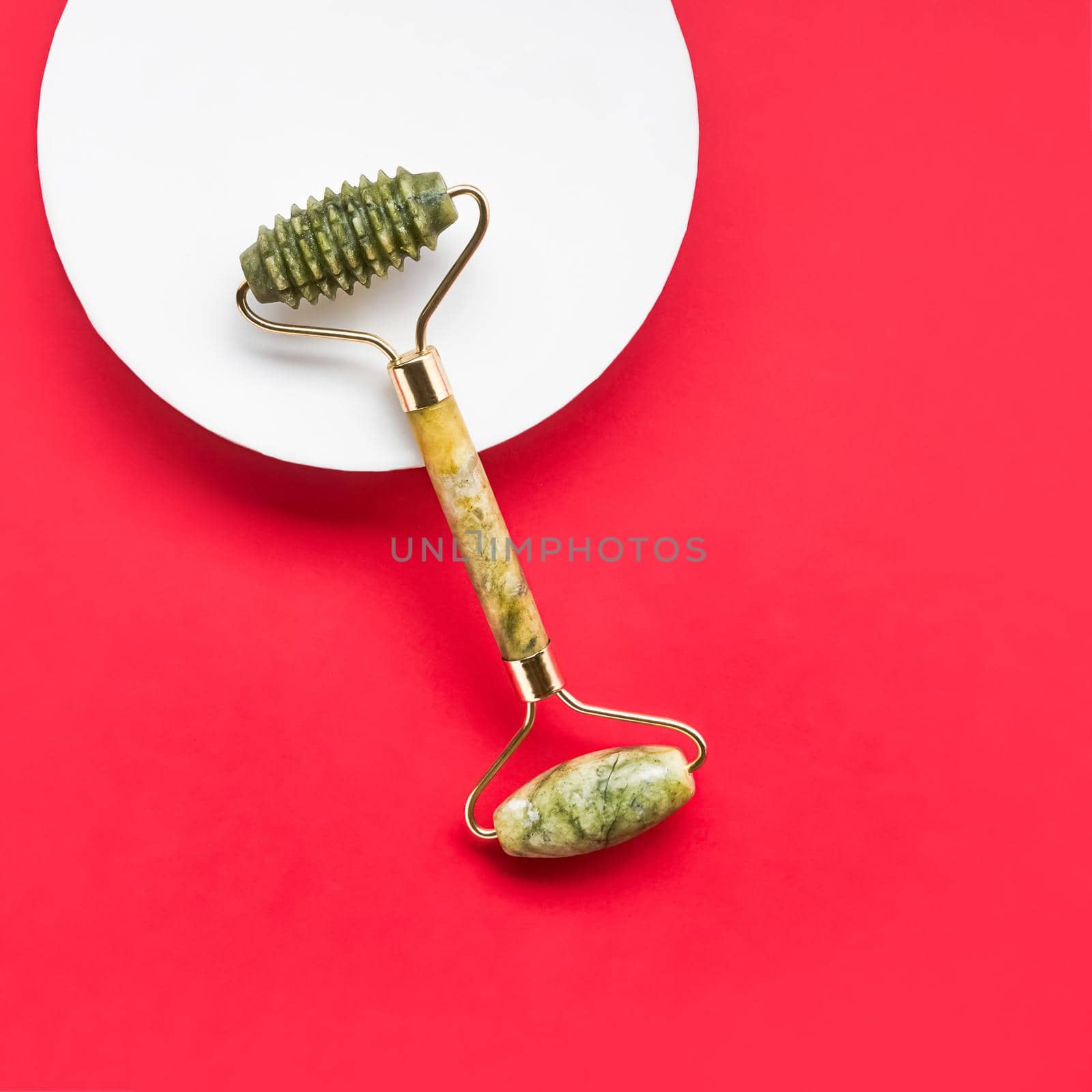 Green face roller from natural jade stone on a red background. SPA concept. Top view, copy space for text