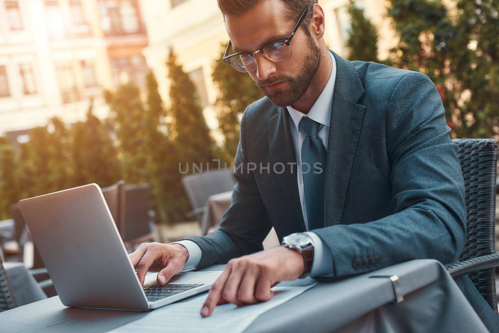 Portrait of handsome bearded businessman in eyeglasses working with laptop while sitting in restaurant outdoors. Digital concept. Business concept