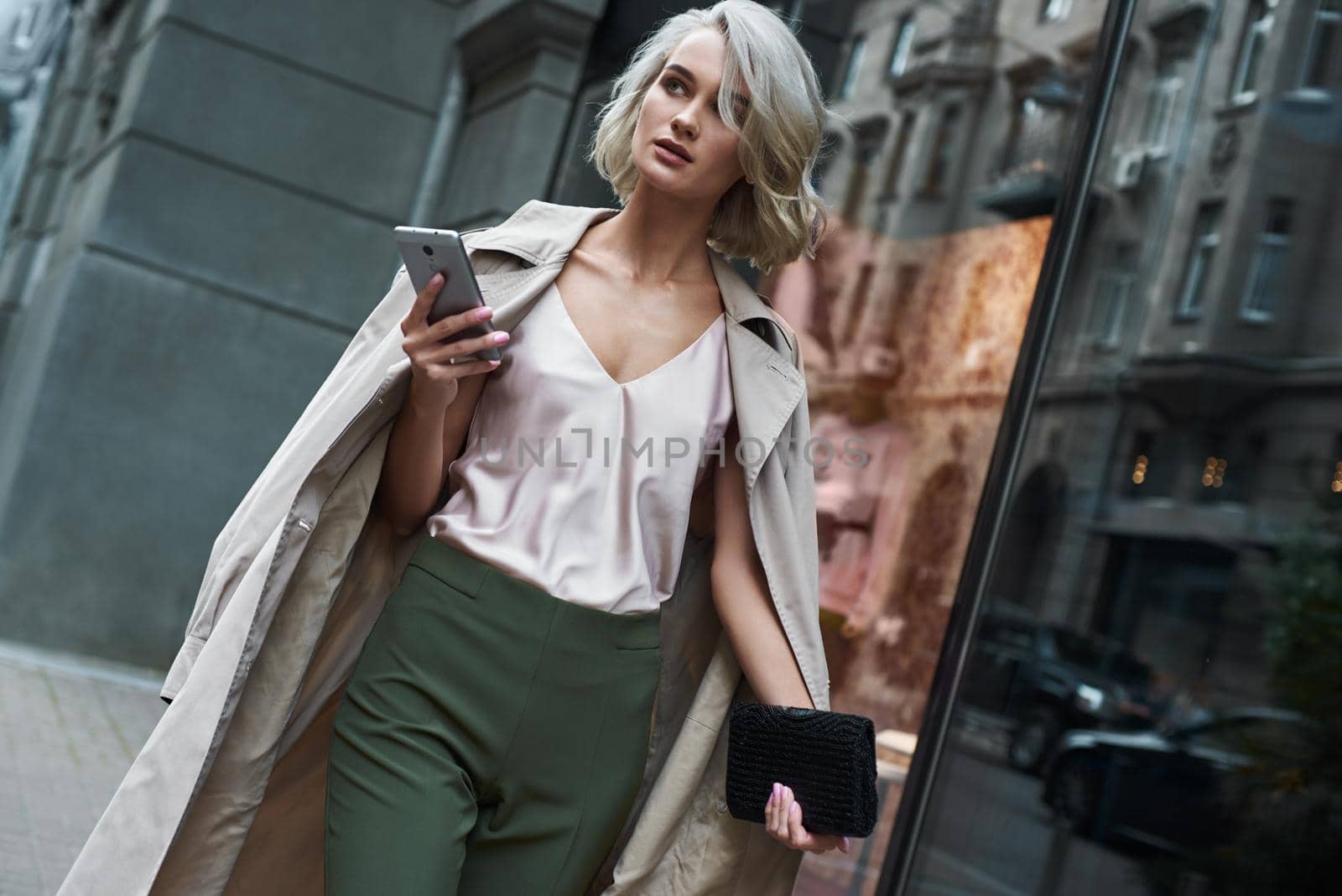 Fashion. Young stylish woman walking on the city street browsing internet on smartphone looking aside pensive