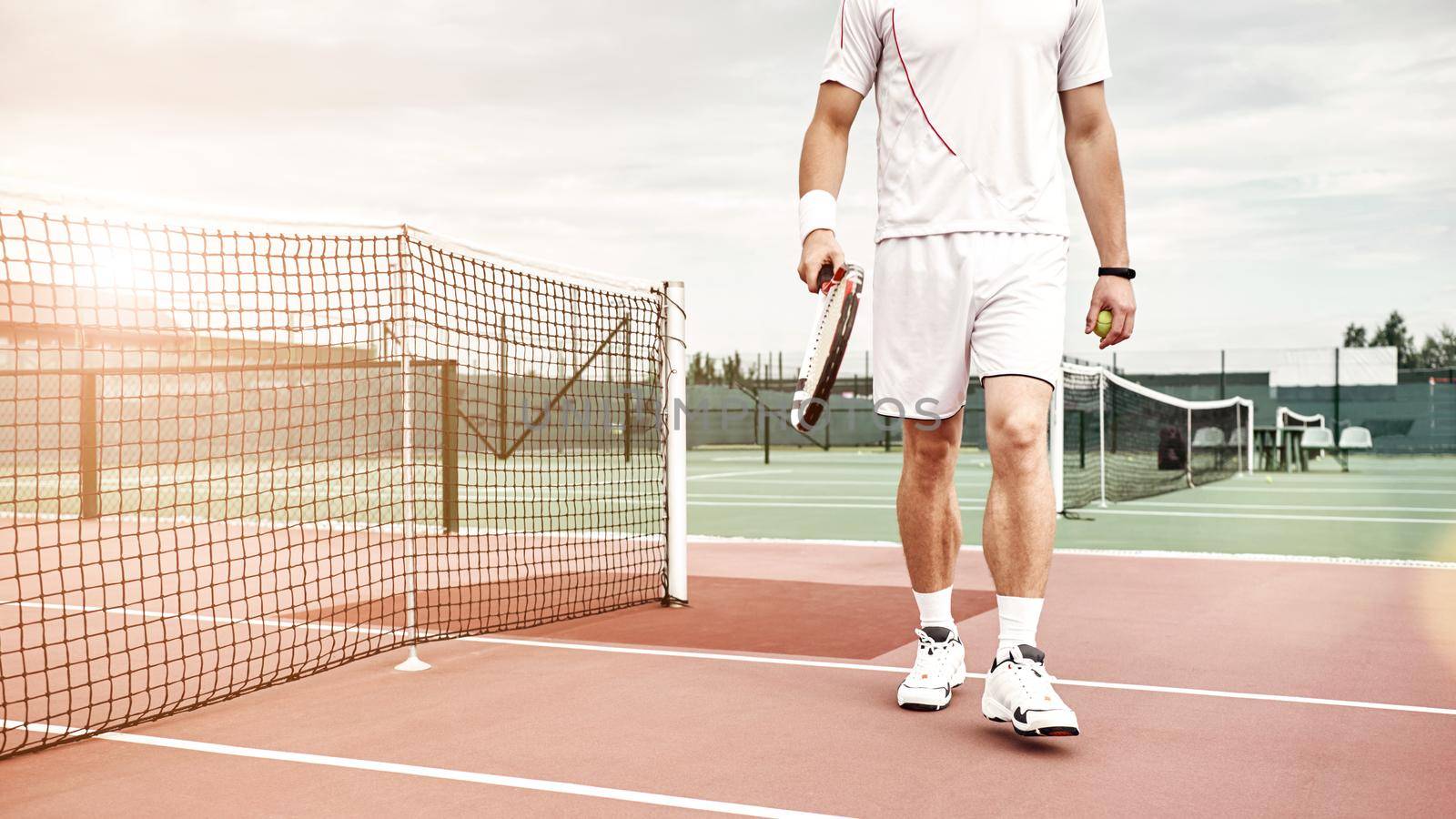 Cropped shot of tennis player in white polo and shorts with the racket and the ball standing on the court