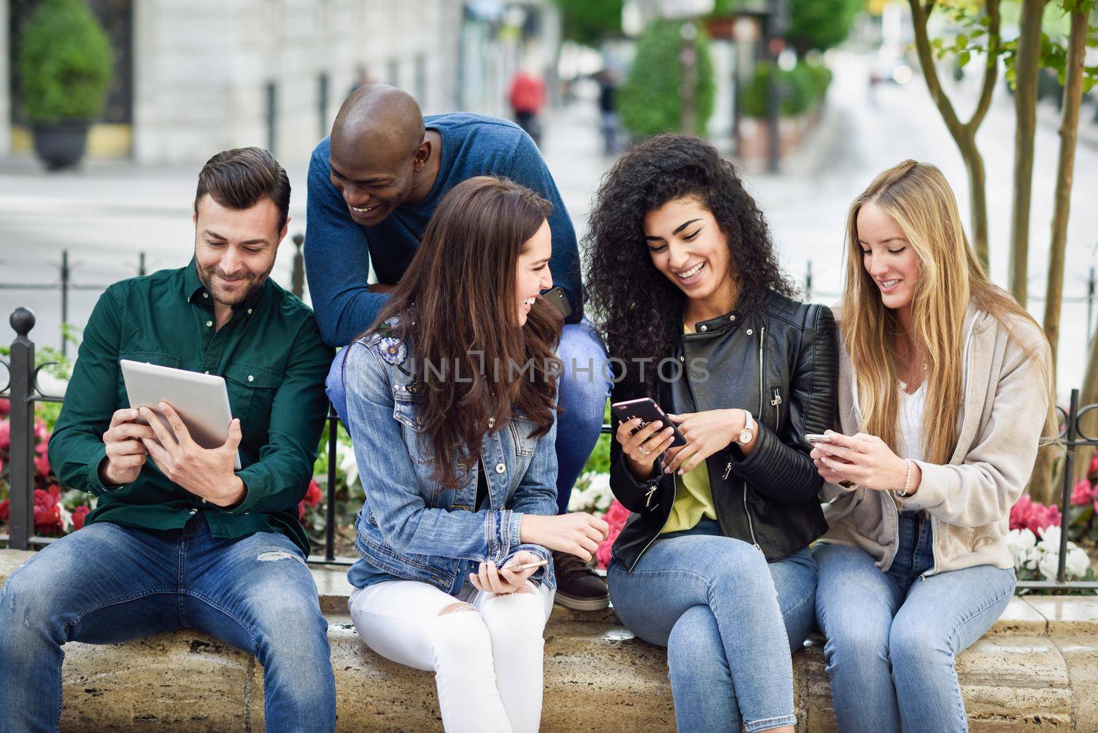 Multi-ethnic group of young people using smartphone and tablet computers outdoors in urban background. Women and men smiling and laughing in the street wearing casual clothes.