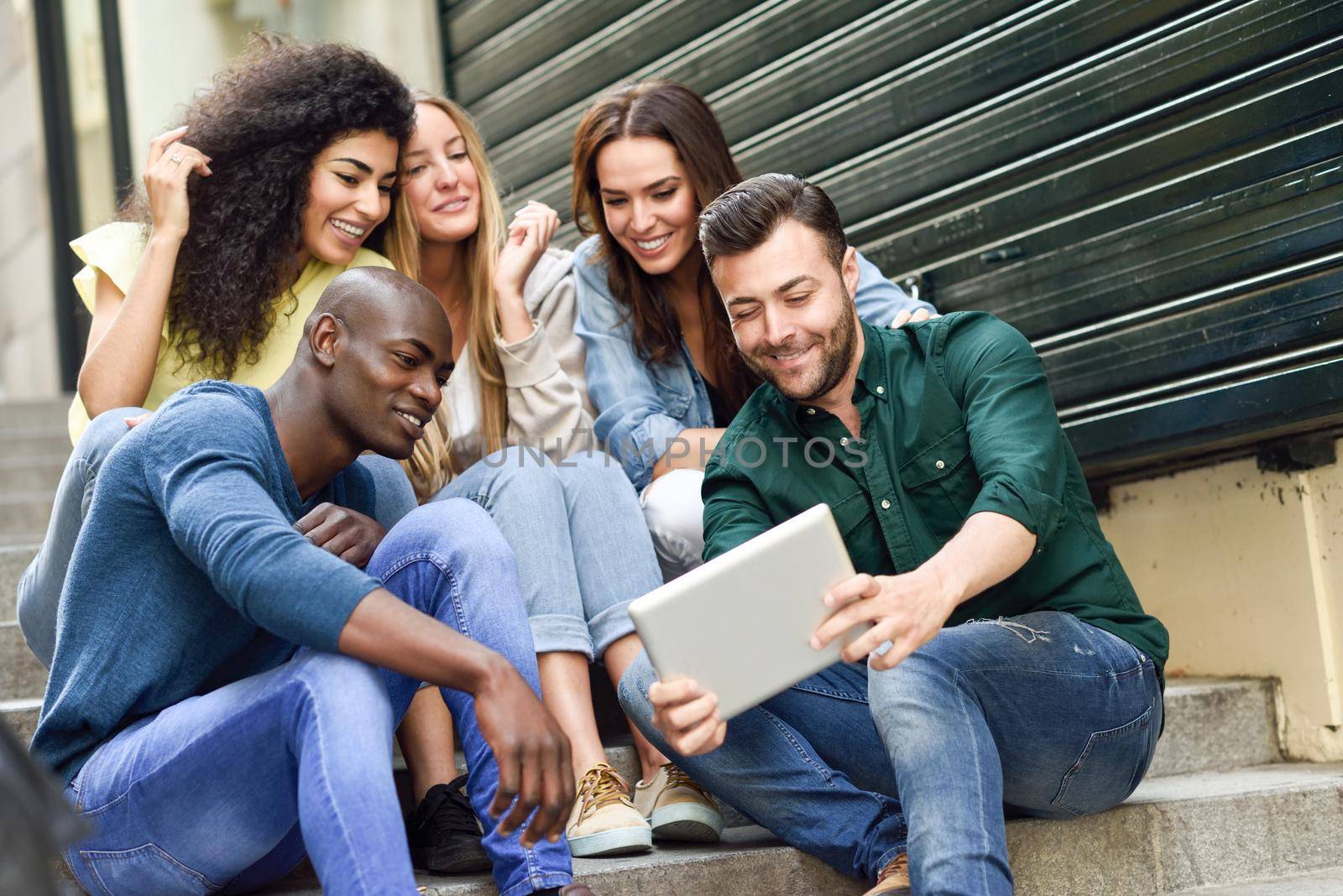 Multi-ethnic group of young people looking at a tablet computer outdoors in urban background. Group of men and woman sitting together on steps.
