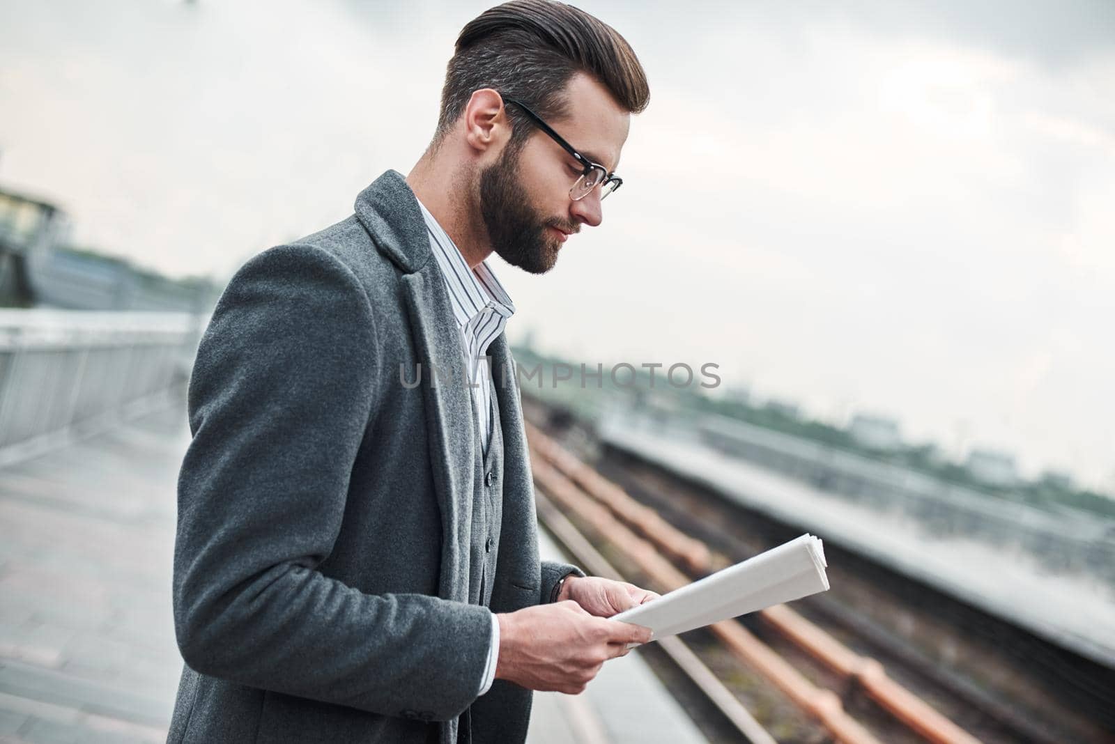 Business trip. Young businessman standing near railway reading newspaper close-up by friendsstock