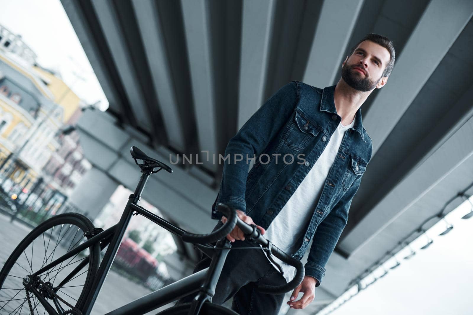 Outdoors leisure. Young stylish man standing on city street with bicycle looking aside smiling confident by friendsstock