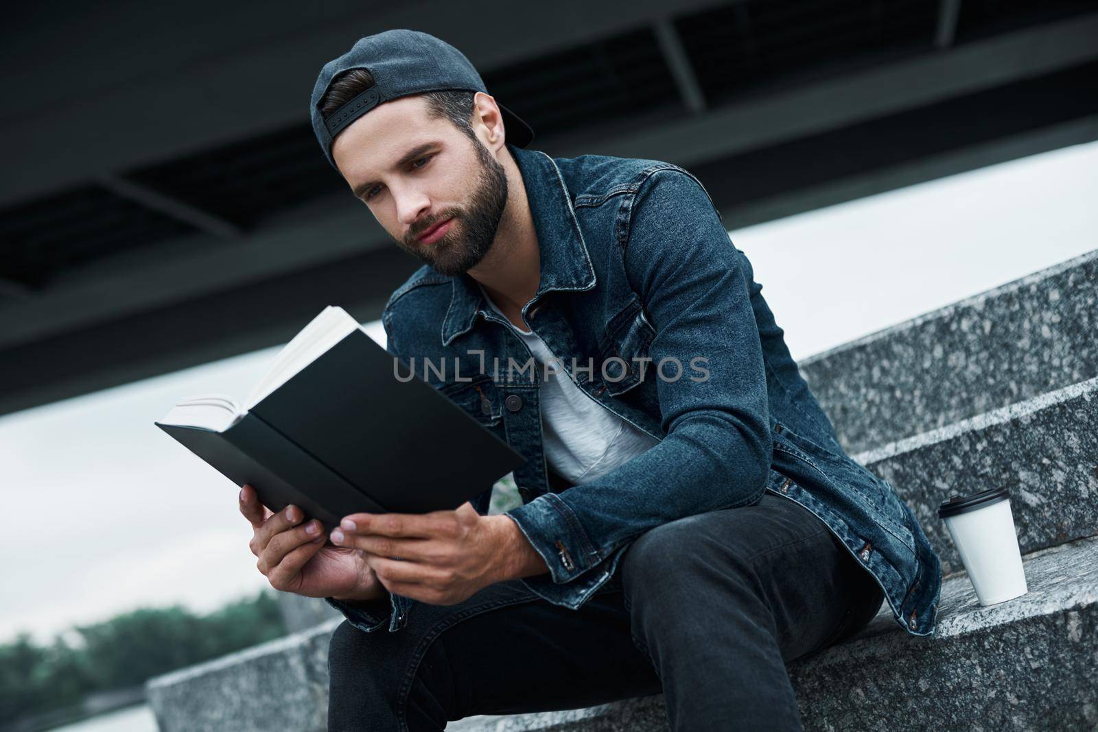 Outdoors leisure. Young stylish man sitting on stairs on city street with cup of hot coffee reading book joyful close-up by friendsstock