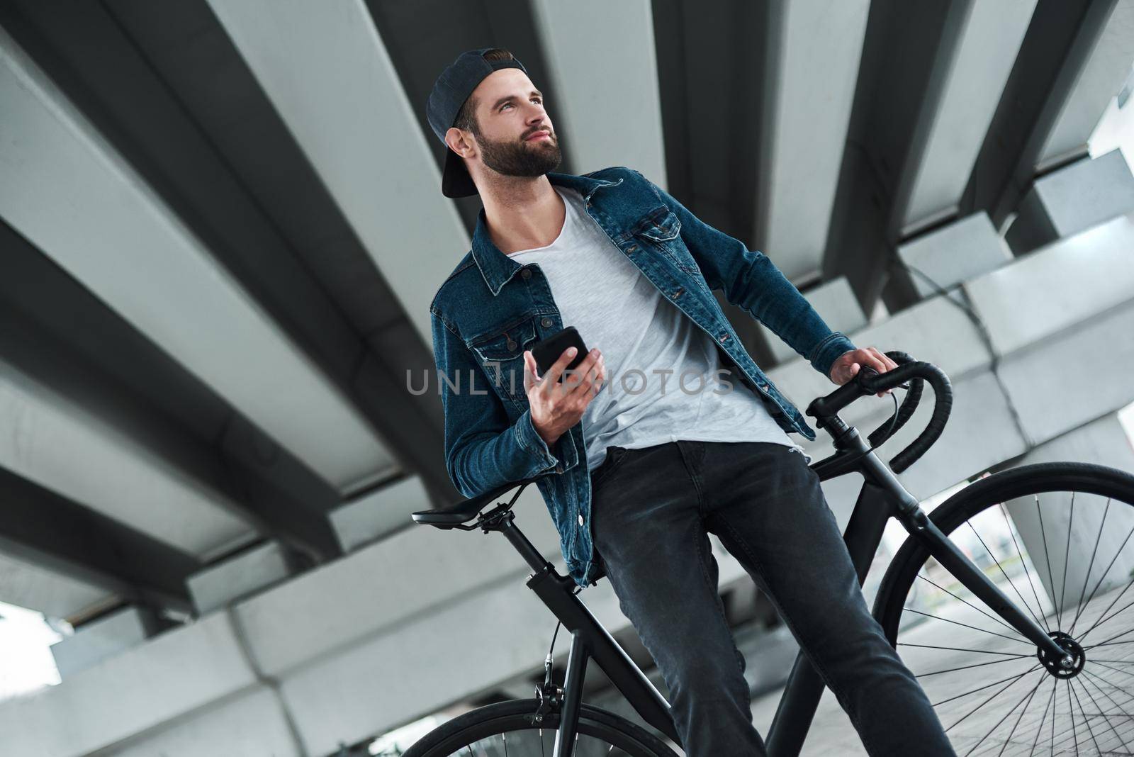 Outdoors leisure. Young stylish man sitting on bycicle on city street holding smartphone looking aside smiling joyful