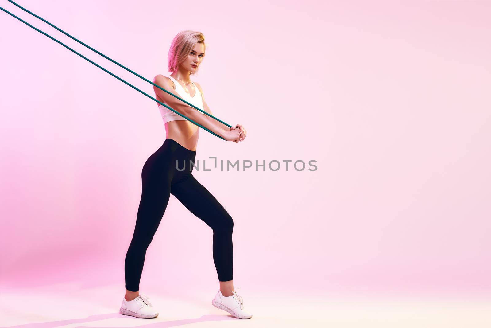 Training body . Side view of sporty young woman in white top and black leggings exercising with resistance band while standing against pink background in studio by friendsstock