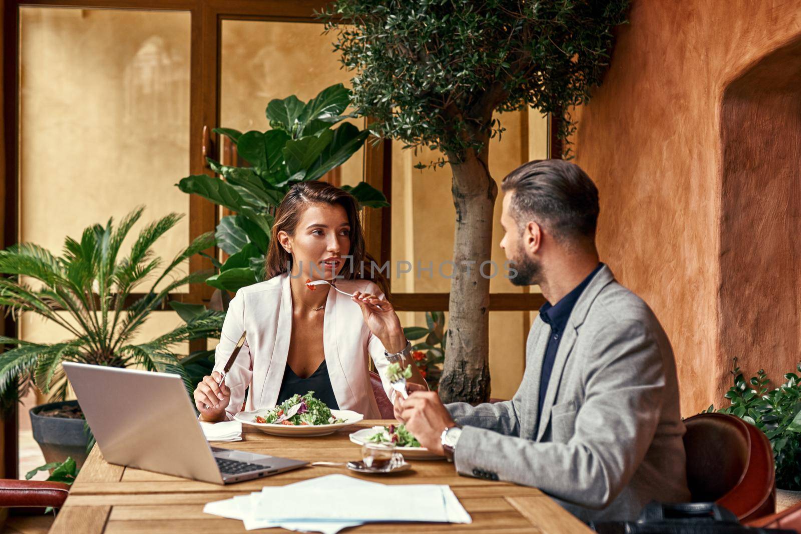 Business lunch. Man and woman sitting at table at restaurant eating healthy fresh salad discussing project by friendsstock