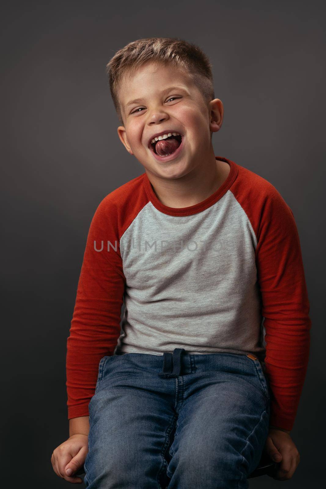 Facial expressions, emotions, feelings. Funny laughing boy in red shirt with eyes shut. Happy kid on dark grey background. Funny family time. Little boy enjoying having fun in studio.