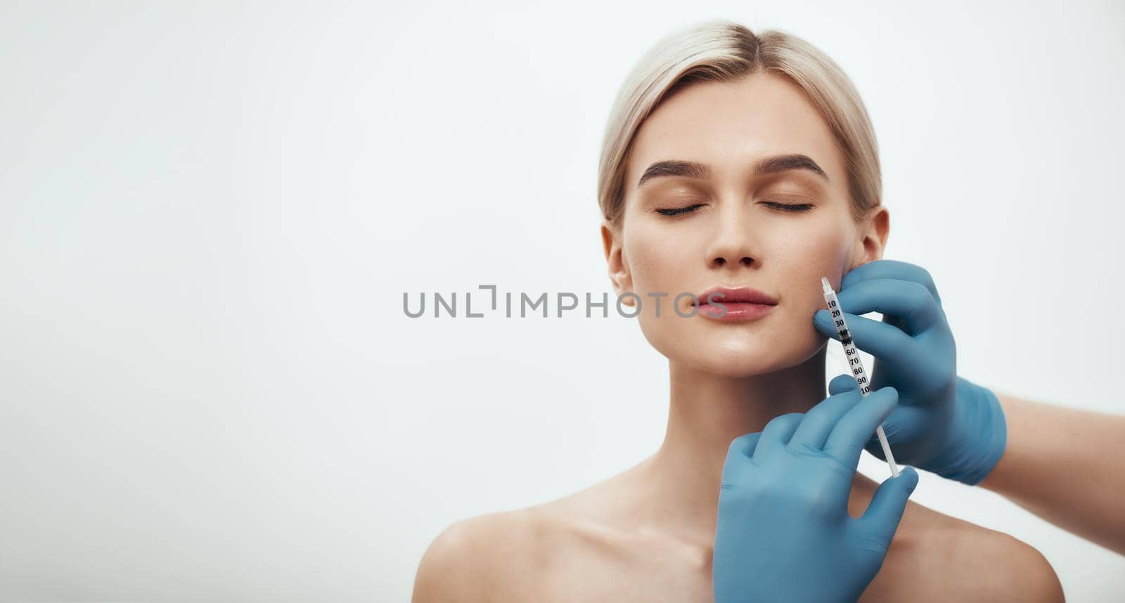 Young pretty woman keeping her eyes closed while doctors hand making an injection in her face. Horizontal web banner. Studio shot. Beauty concept