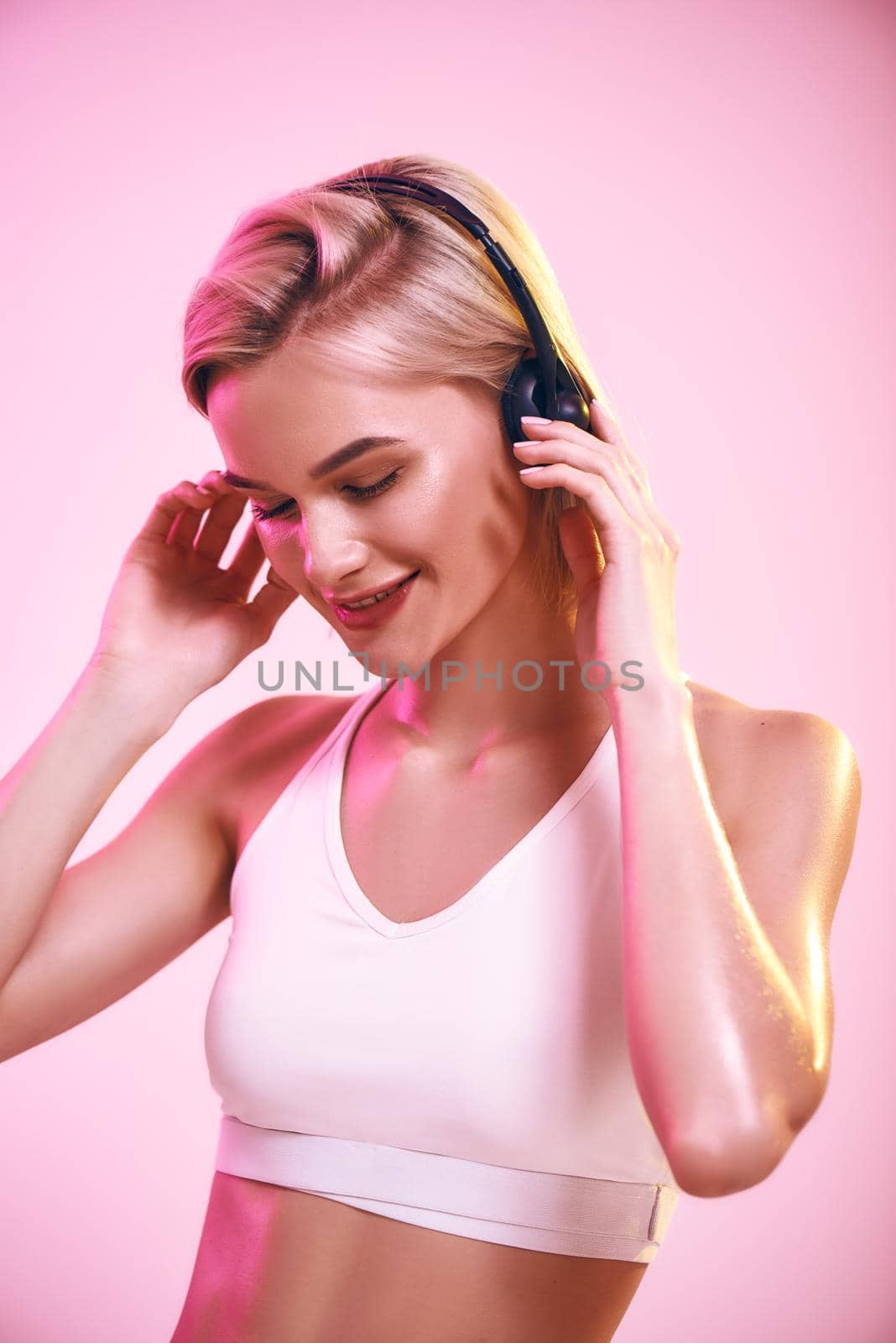 Enjoying music. Close up of cute and sporty woman in headphones listening music and smiling while standing against pink background by friendsstock