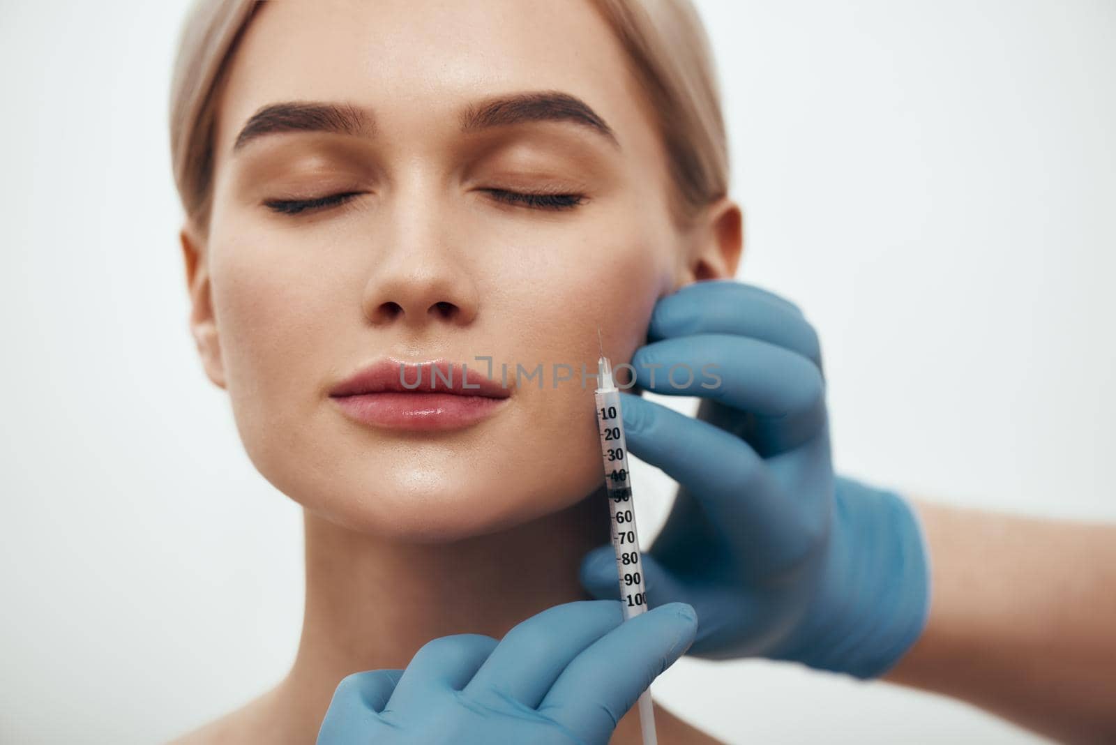 Wanna be perfect. Portrait of young pretty woman keeping eyes closed while doctors hand in blue medical gloves making an injection in her face by friendsstock