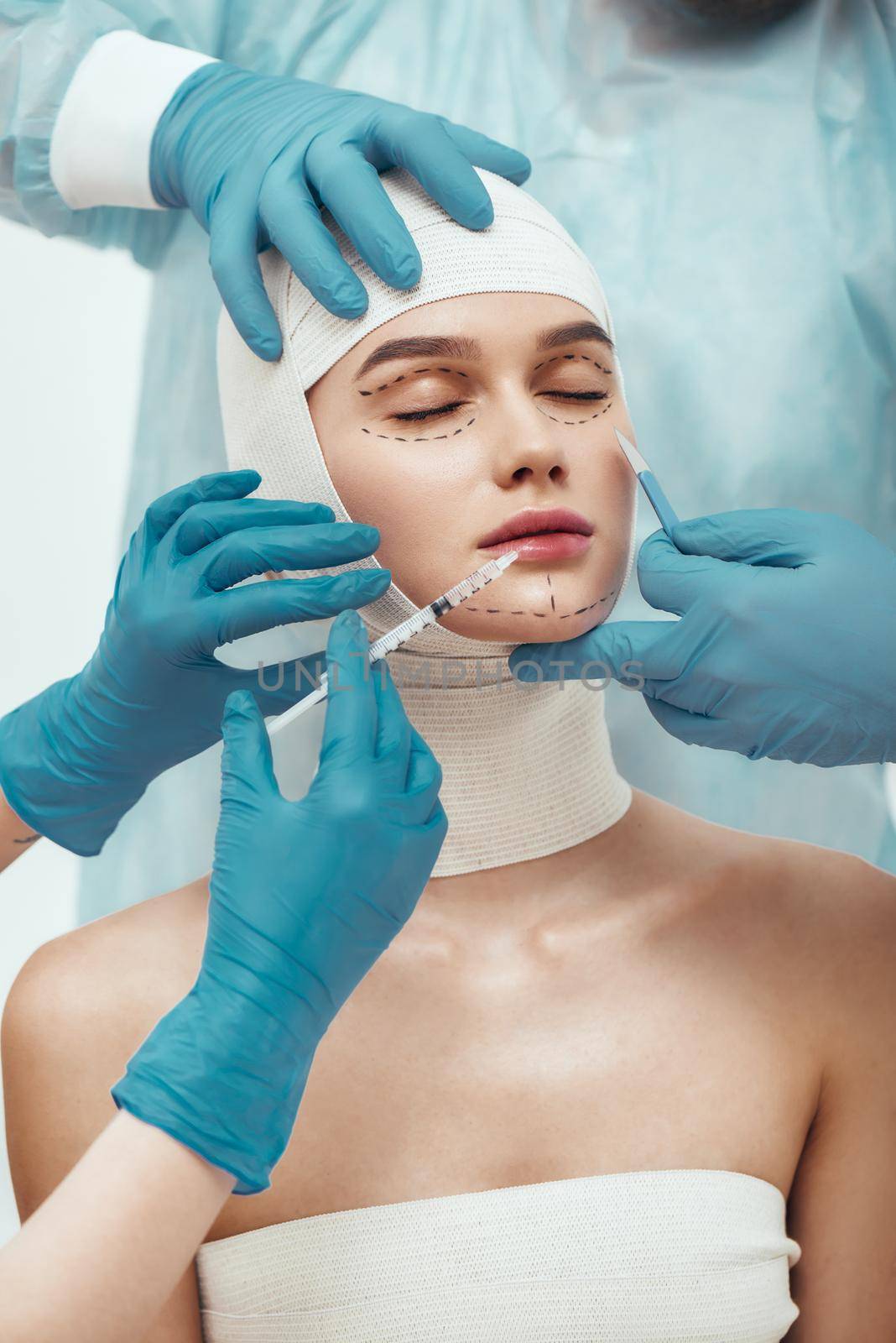 No chance for wrinkles. Young woman with head in bandages keeping eyes closed while plastic surgeons in blue gloves are working with her face. Doctors holding scalpel and syringe near patient face by friendsstock