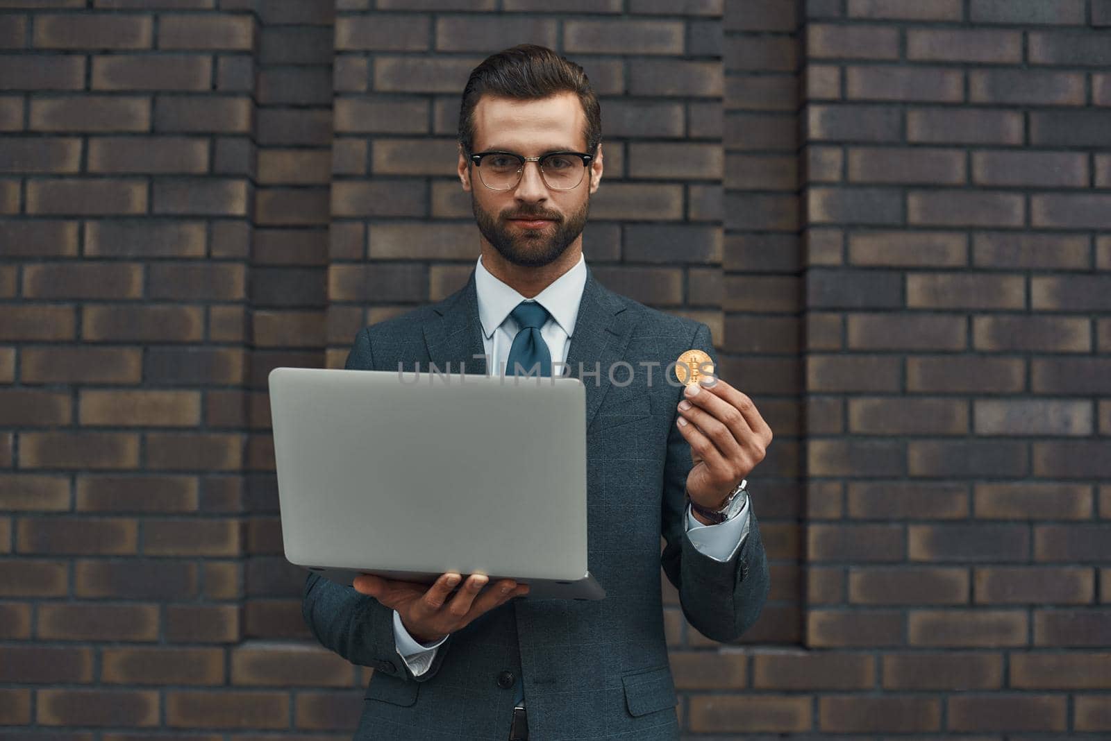 Our future. Handsome businessman in formal wear and eyeglasses holding laptop and bitcoin in another hand while standing against brick wall by friendsstock