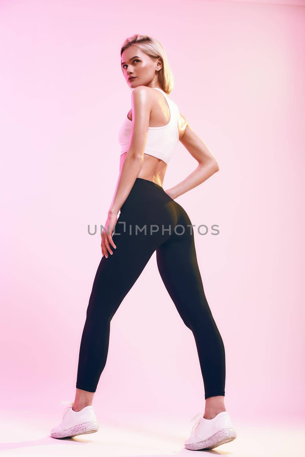 Perfect buttocks. Back view of sporty and cute slim woman with ideal body in sportswear looking at camera while standing against pink background in studio by friendsstock