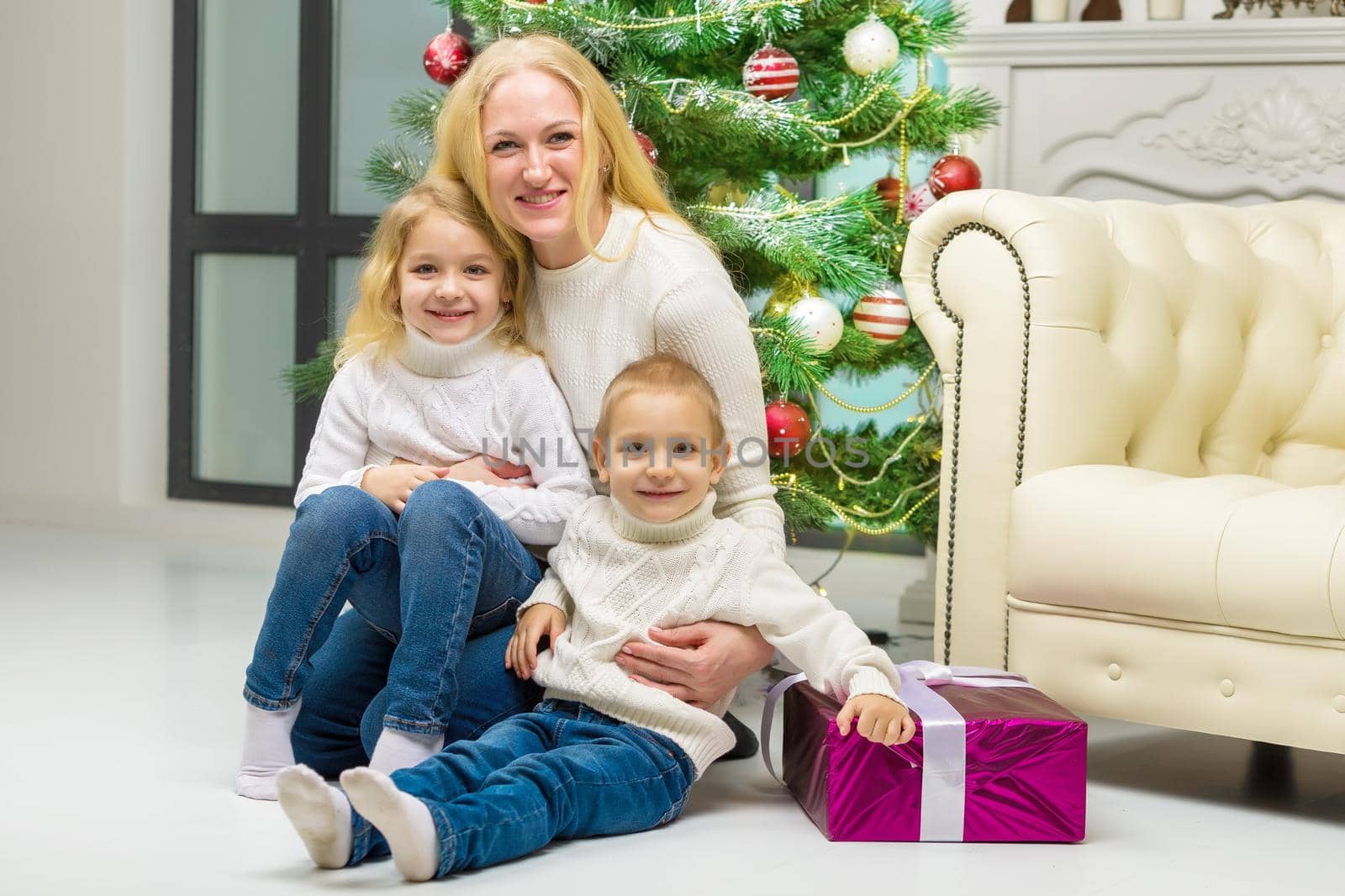 Cheerful Mom Hugging her Cute Little Daughter and Son Happy Family Sitting on the Floor, Smiling Parent and Little Kids Sitting Next Decorated Christmas Tree and Looking at Camera