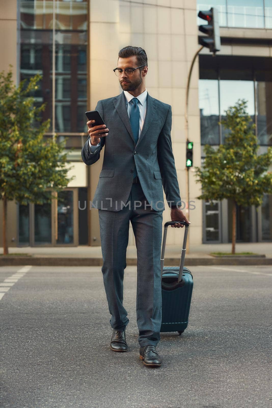 Always in touch. Full length of young and handsome bearded man in suit pulling his luggage and looking at his smartphone while walking outdoors. Travelling. Work concept