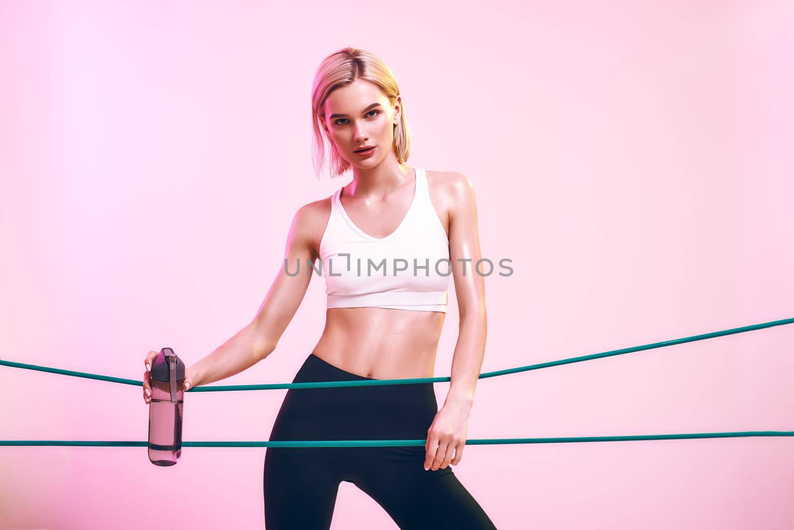 Fitness girl. Sporty young woman in white top and black leggings holding bottle of water and working out with elastic band while standing against pink background in studio. Sport concept. Sport equipment