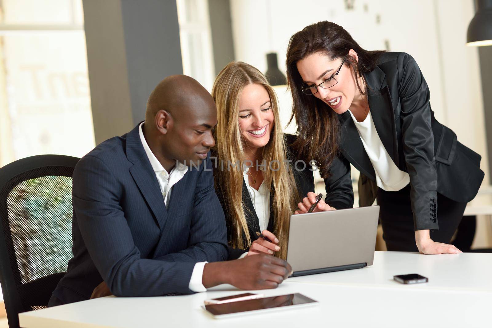 Multi-ethnic group of three businesspeople meeting in a modern office. Two caucasian women and a black man wearing suit looking at a laptop computer.