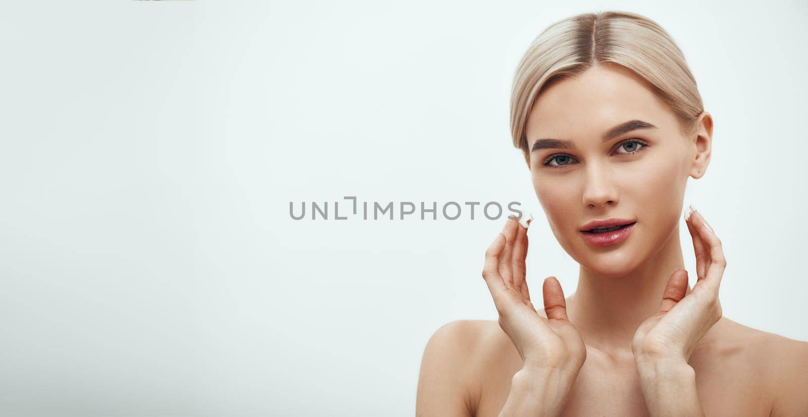 Horizontal web banner. Beautiful young blonde woman touching her face and looking at camera while standing against grey background. Skin care. Beauty concept