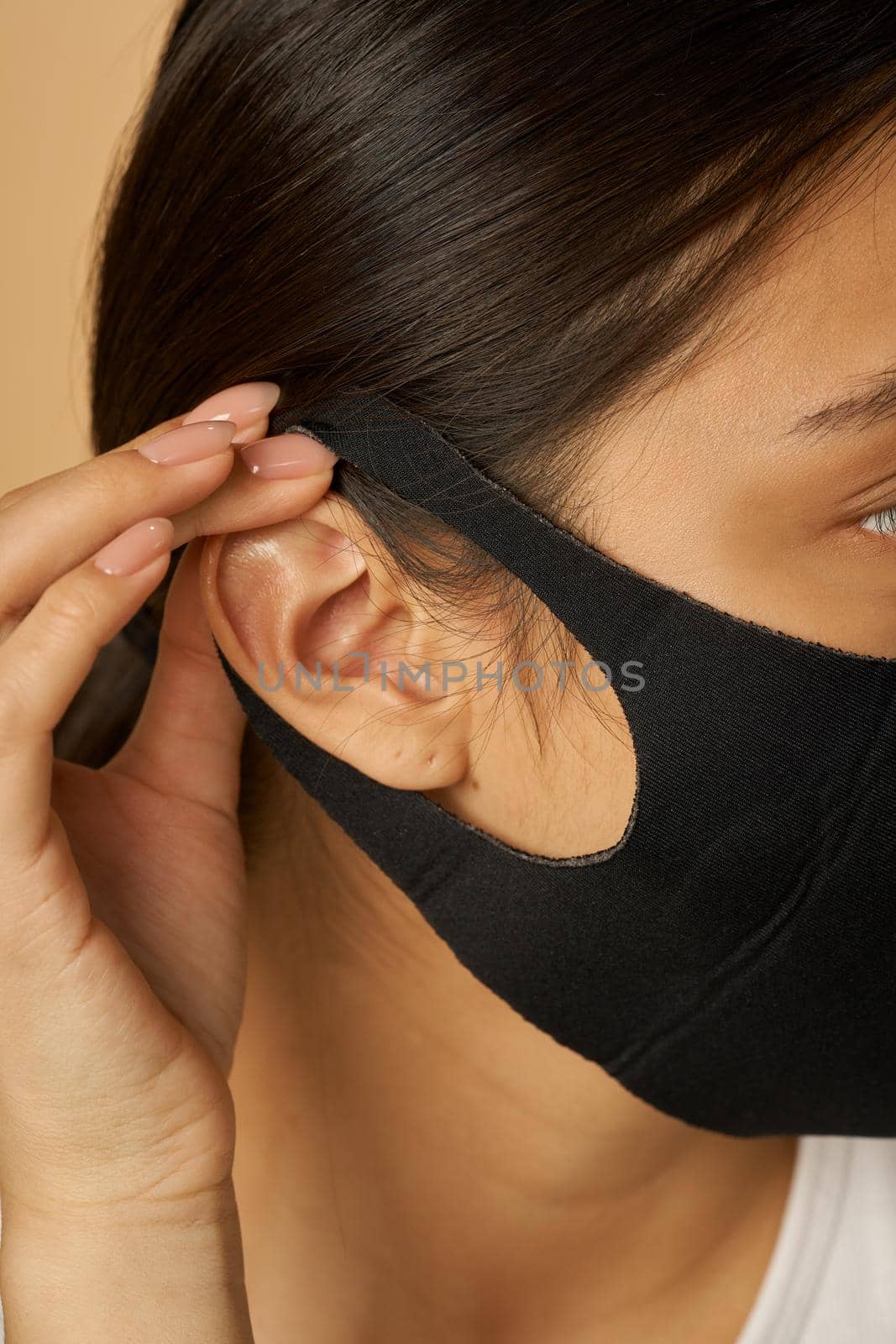 Close up shot of young woman putting on black facial mask, staying safe while posing isolated over beige background. Safety, pandemic concept