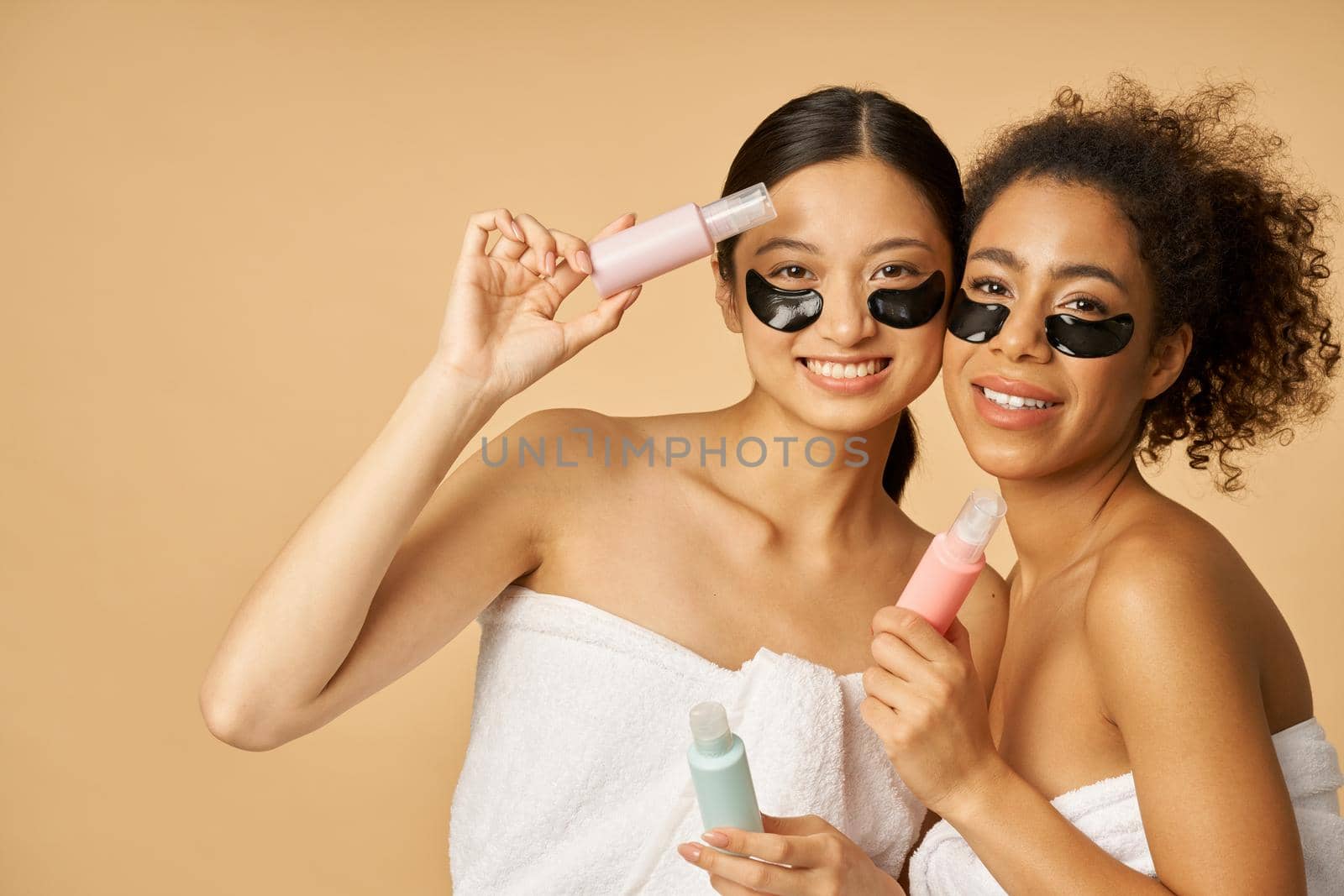 Two joyful young women with applied hydro gel under eye patches, wrapped in towel holding bottles with beauty product, posing isolated over beige background by friendsstock