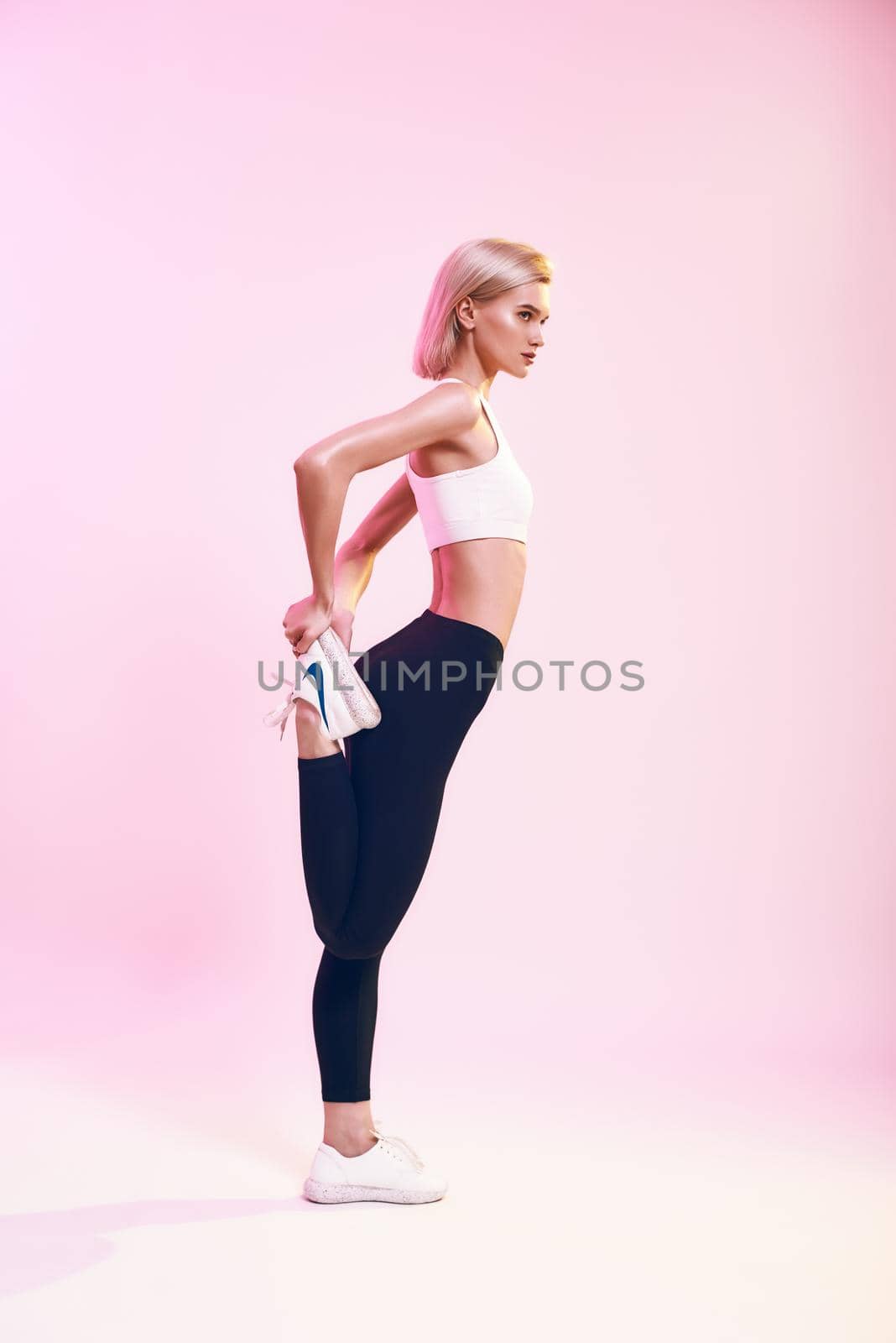 Strong and flexible. Back view of sporty and cute slim woman in sportswear stretching her legs while standing against pink background in studio. Sport. Active lifestyle. Workout