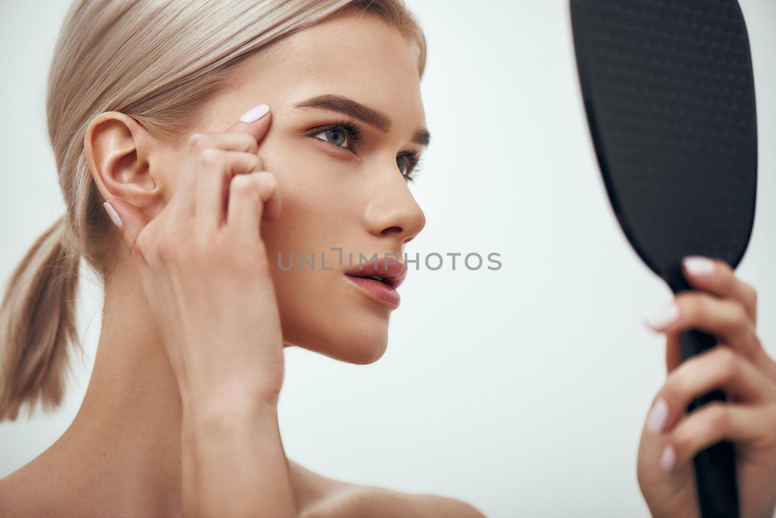 Mimic wrinkles. Profile portrait of young attractive woman touching her face and looking in small mirror while standing against grey background by friendsstock