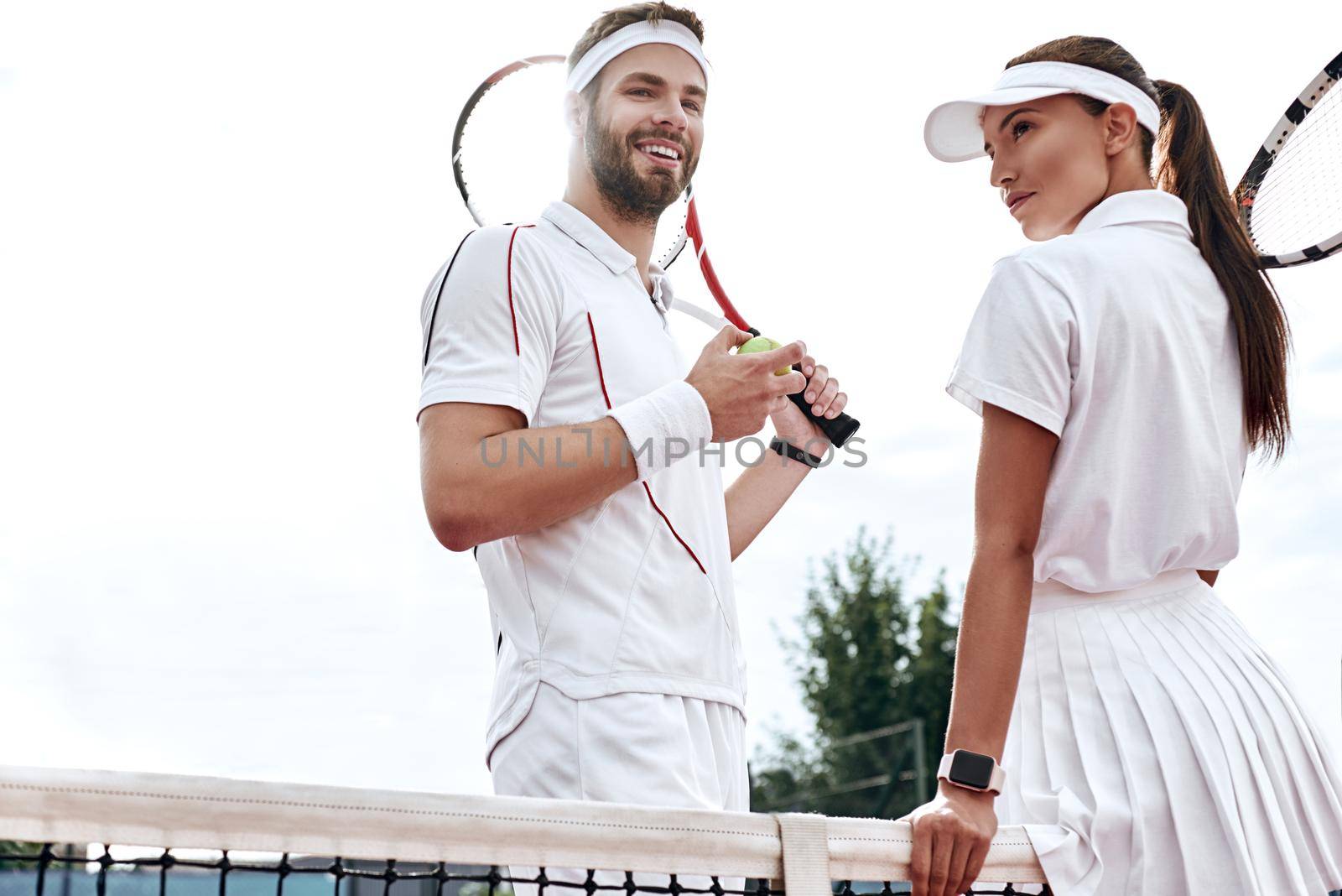 Beautiful woman and handsome man are going to play tennis. Woman in white polo and skirt is looking away. Man in white sportswear with the racket and the ball in his hands smiles