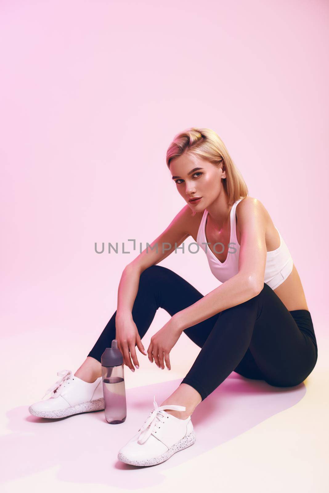 After workout. Cute and young sporty woman in sports clothing relaxing and looking at camera while sitting on the floor against pink background in studio. Sport. Active lifestyle