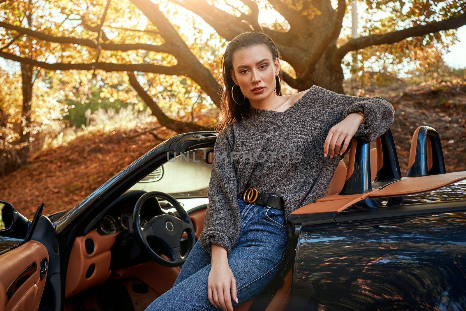 Sexy young woman in jeans and in a grey sweater sits in front seat of a cabriolet. Autumn season