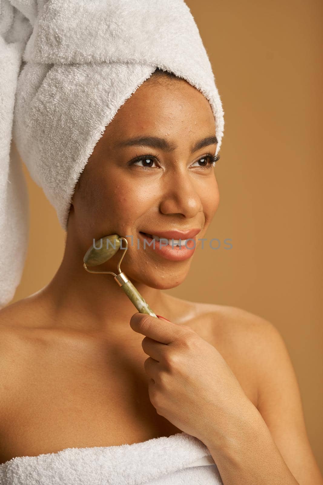 Portrait of cheerful mixed race young woman after shower smiling away while using jade roller for massaging her face isolated over beige background by friendsstock