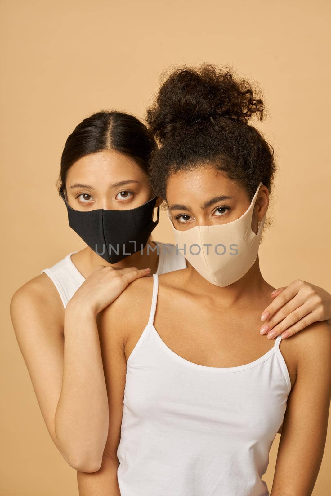 Portrait of two beautiful young diverse women wearing protective facial masks looking at camera while posing together isolated over beige background by friendsstock