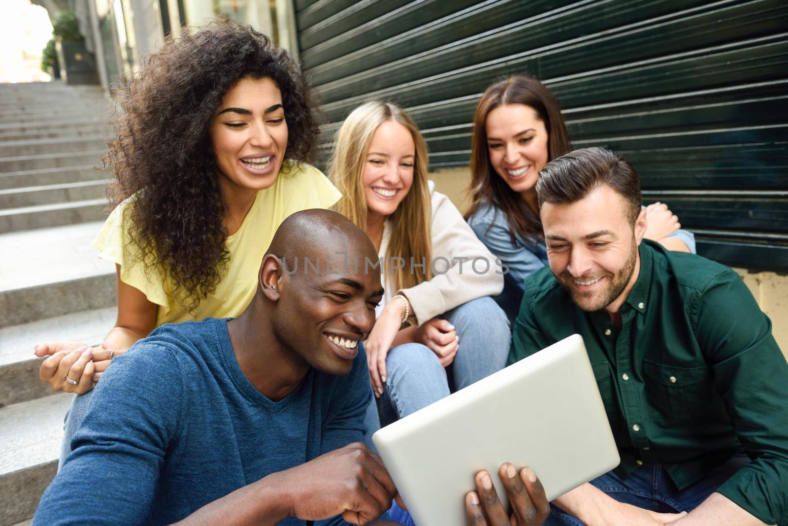 Multi-ethnic group of young people looking at a tablet computer outdoors in urban background. Group of men and woman sitting together on steps.