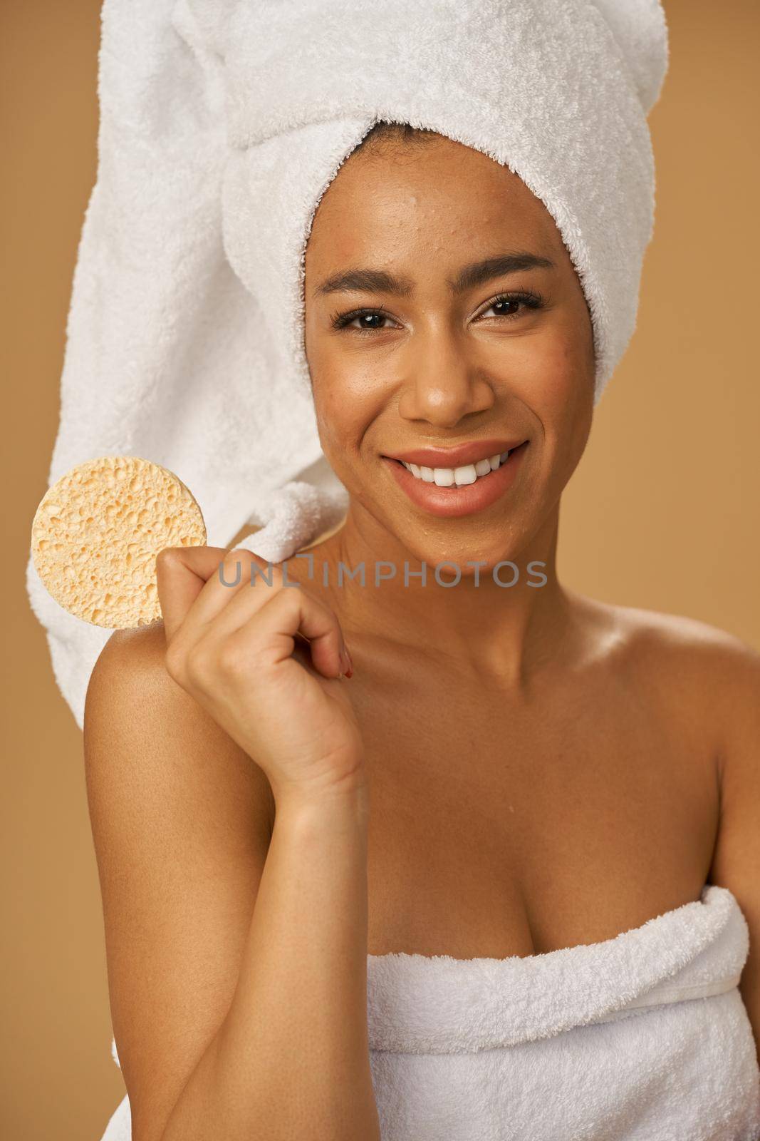 Funny mixed race young woman smiling at camera, holding cleansing face sponge, posing isolated over beige background. Bathroom routine by friendsstock