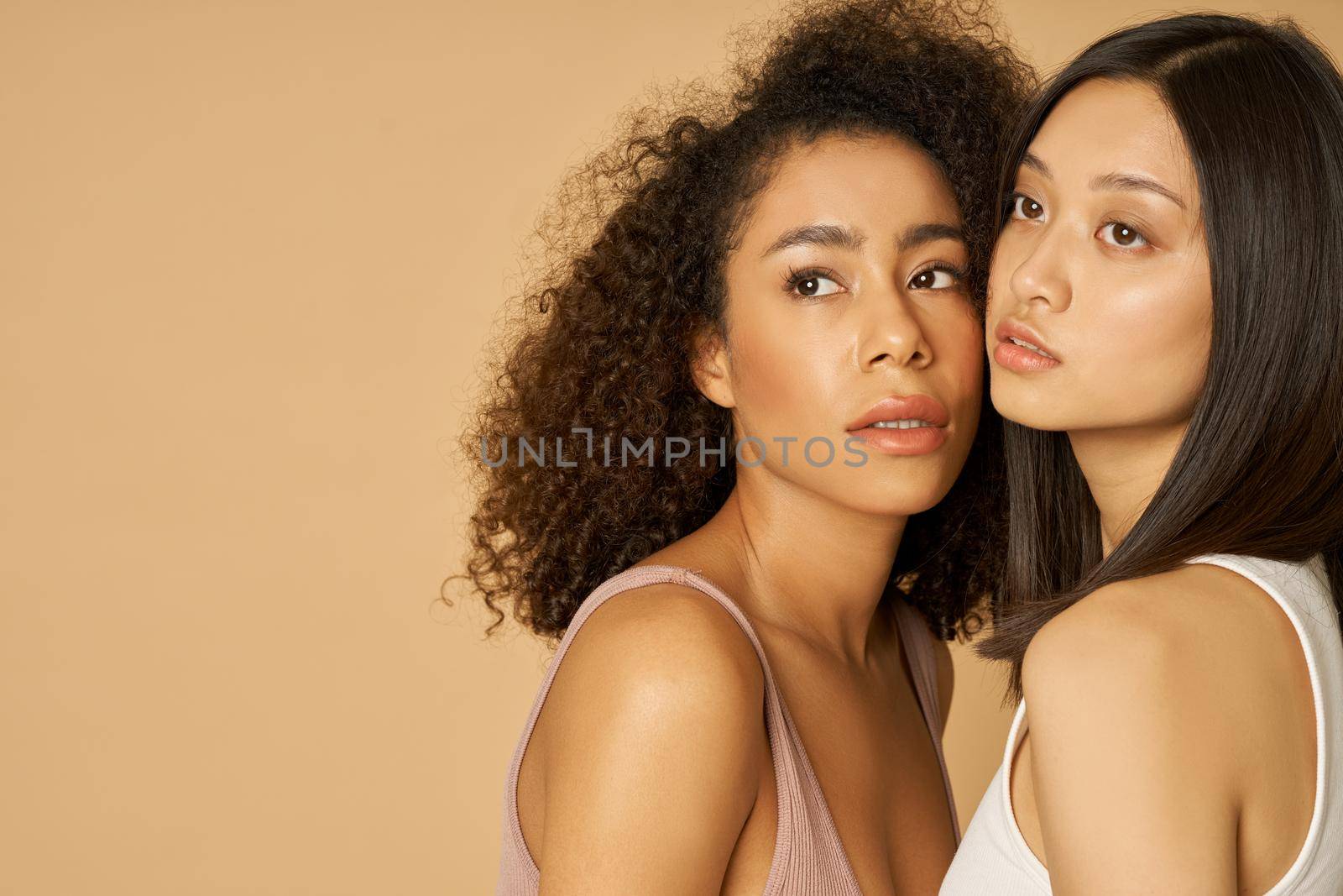 Portrait of two gorgeous mixed race young women with perfect skin looking away while posing isolated over beige background by friendsstock