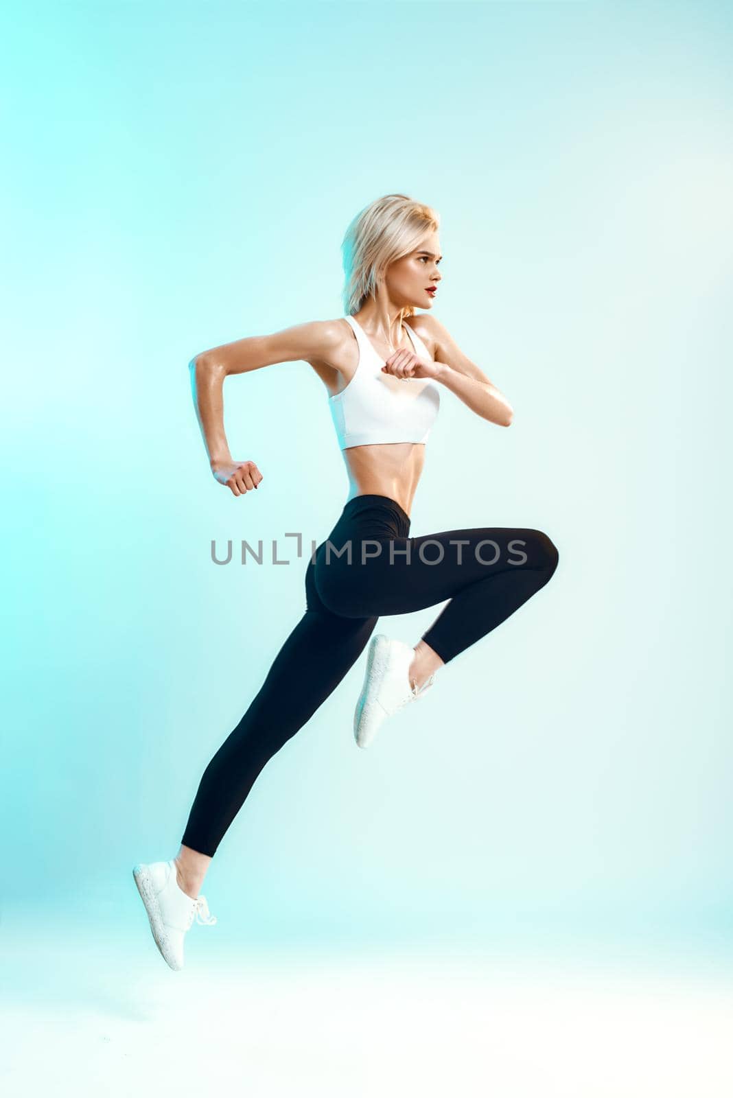 Winner. Side view of sporty young woman in white top and black leggings jumping against blue background in studio. Sport. Active lifestyle. Studio shot