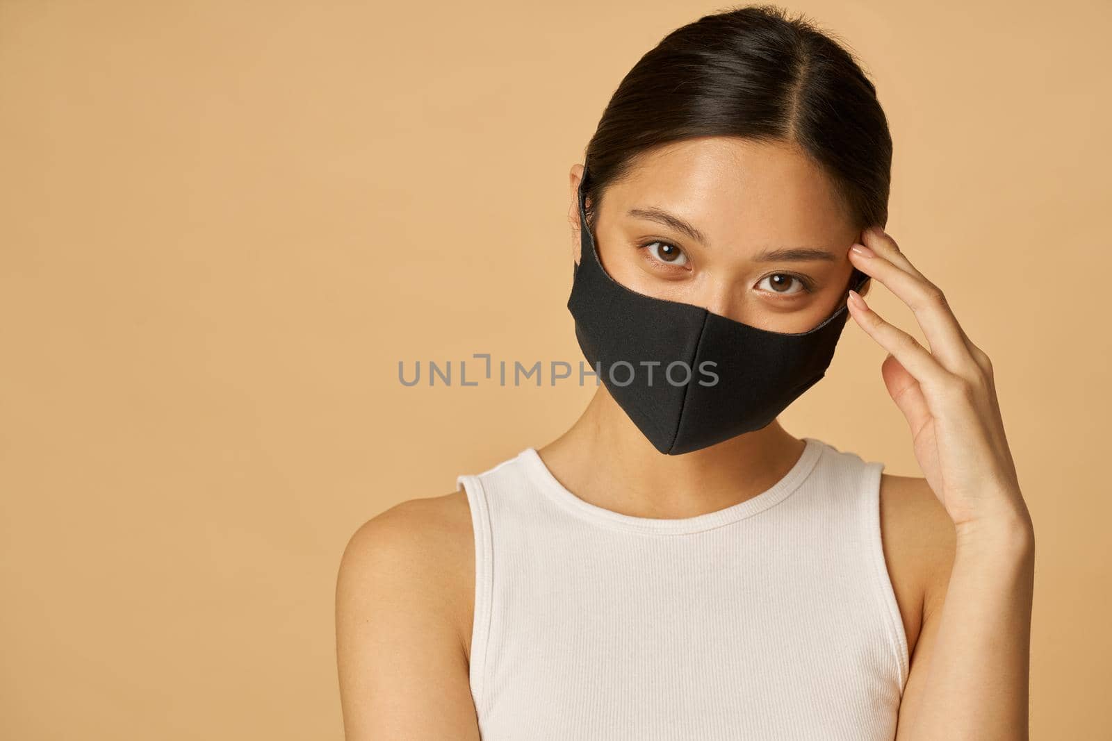 Confident young woman wearing black facial mask looking at camera, posing isolated over beige background. Safety, pandemic concept