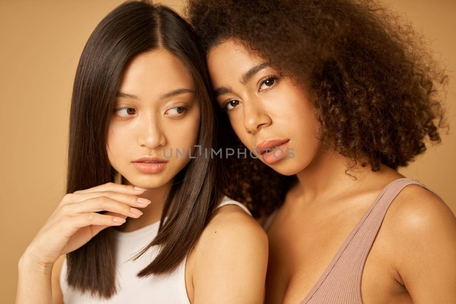 Face closeup of two beautiful mixed race young women with perfect skin posing for camera while standing isolated over beige background. Female beauty portrait