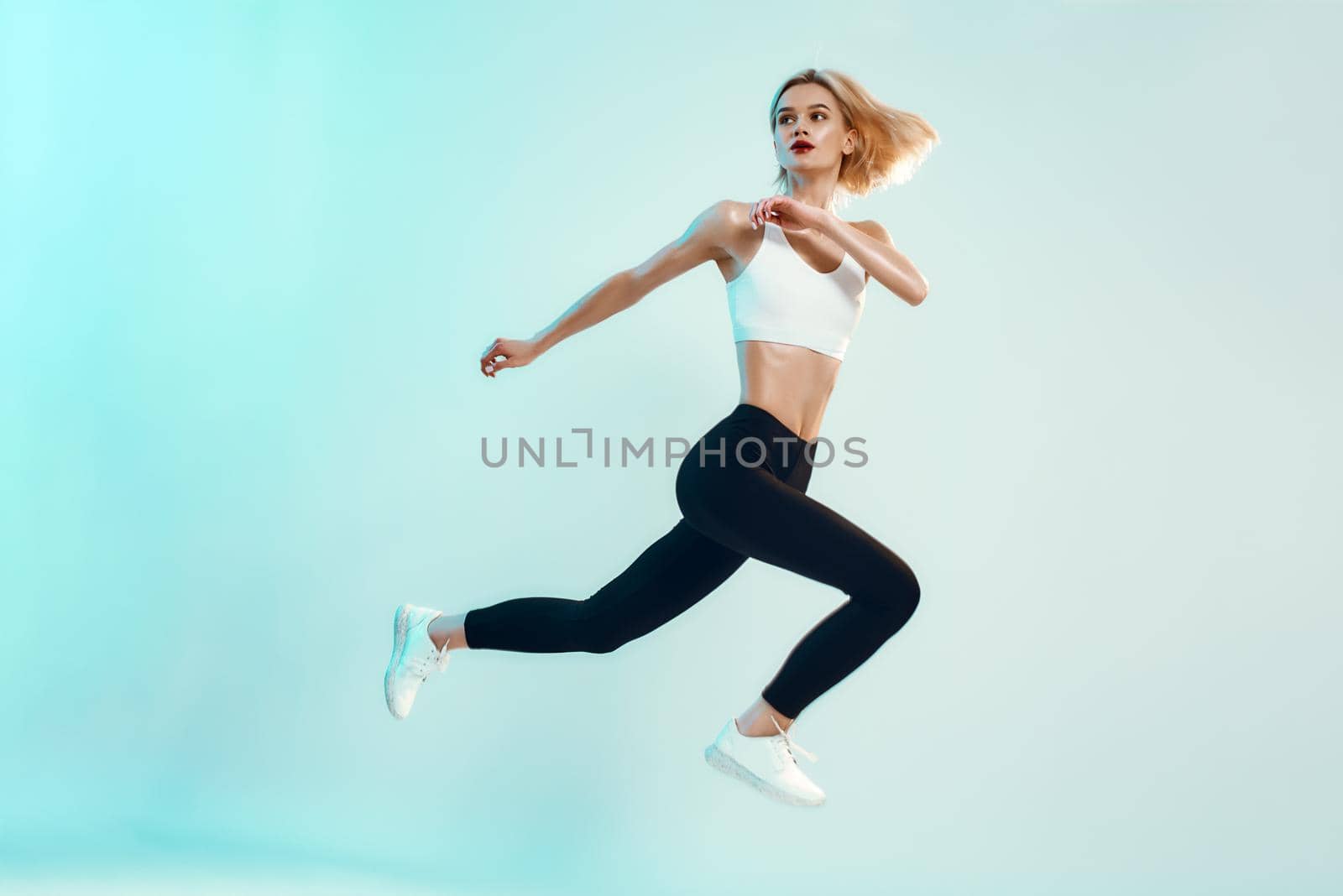 No limits Sporty and young woman with perfect body in white top and black leggings jumping against blue background in studio by friendsstock