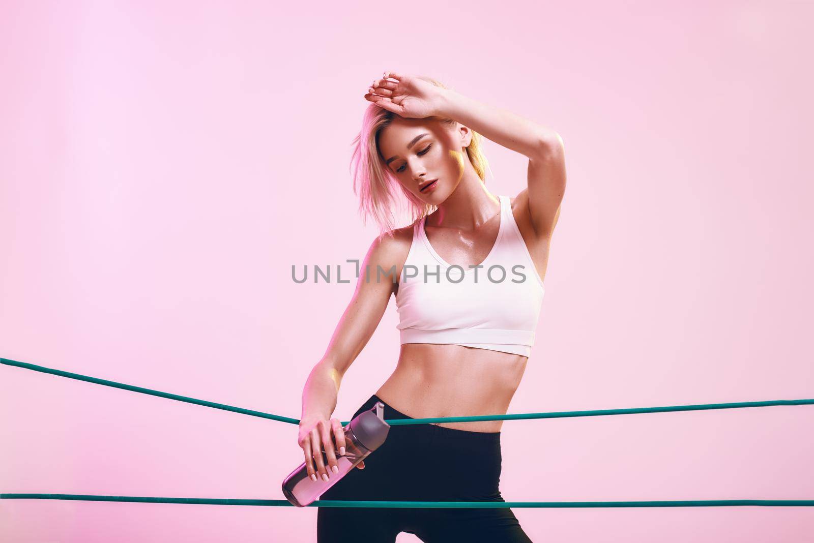 Fit and healthy. Sporty young woman in white top and black leggings holding bottle of water and working out with elastic band while standing against pink background in studio. Sport concept. Sport equipment