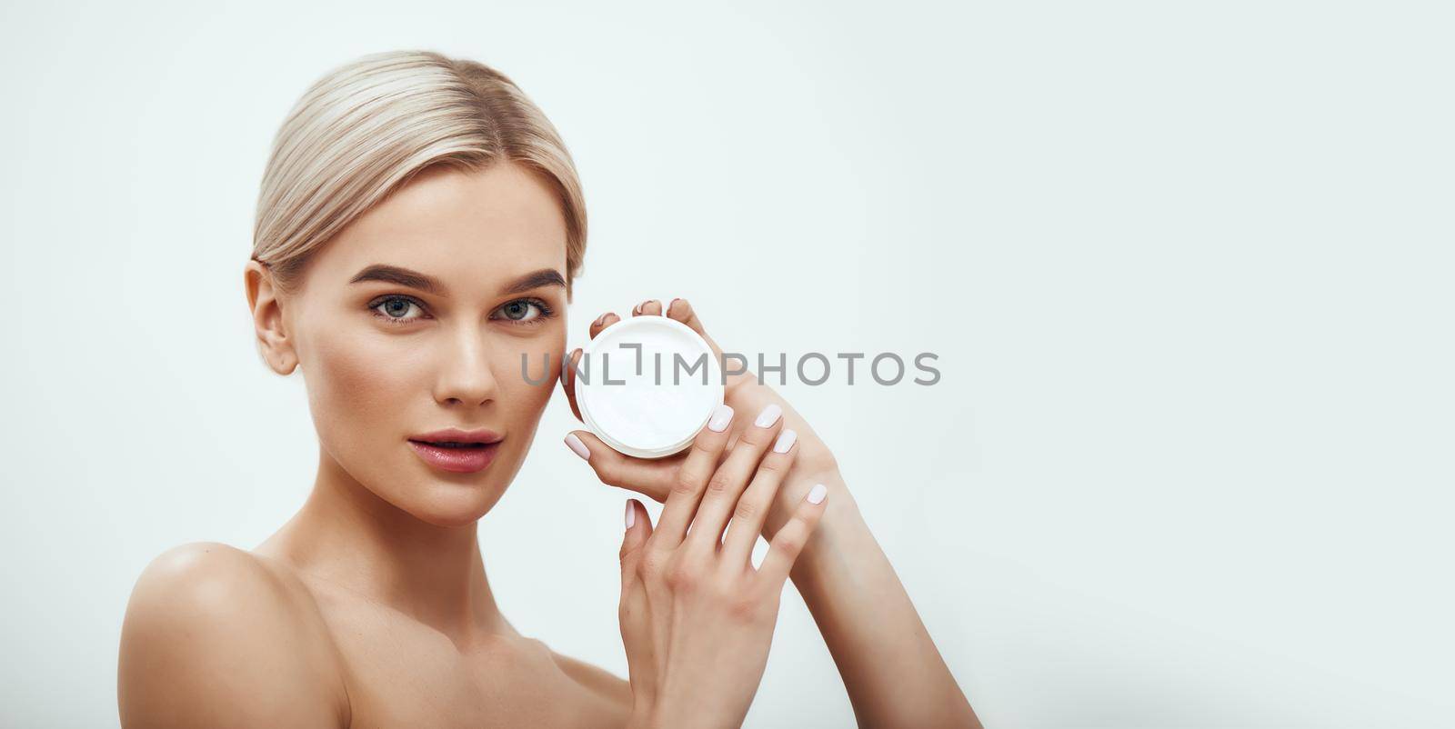 You need a special face cream. Cute young blonde woman showing face cream while standing against grey background. Skin care. Horizontal web banner. Cosmetics concept