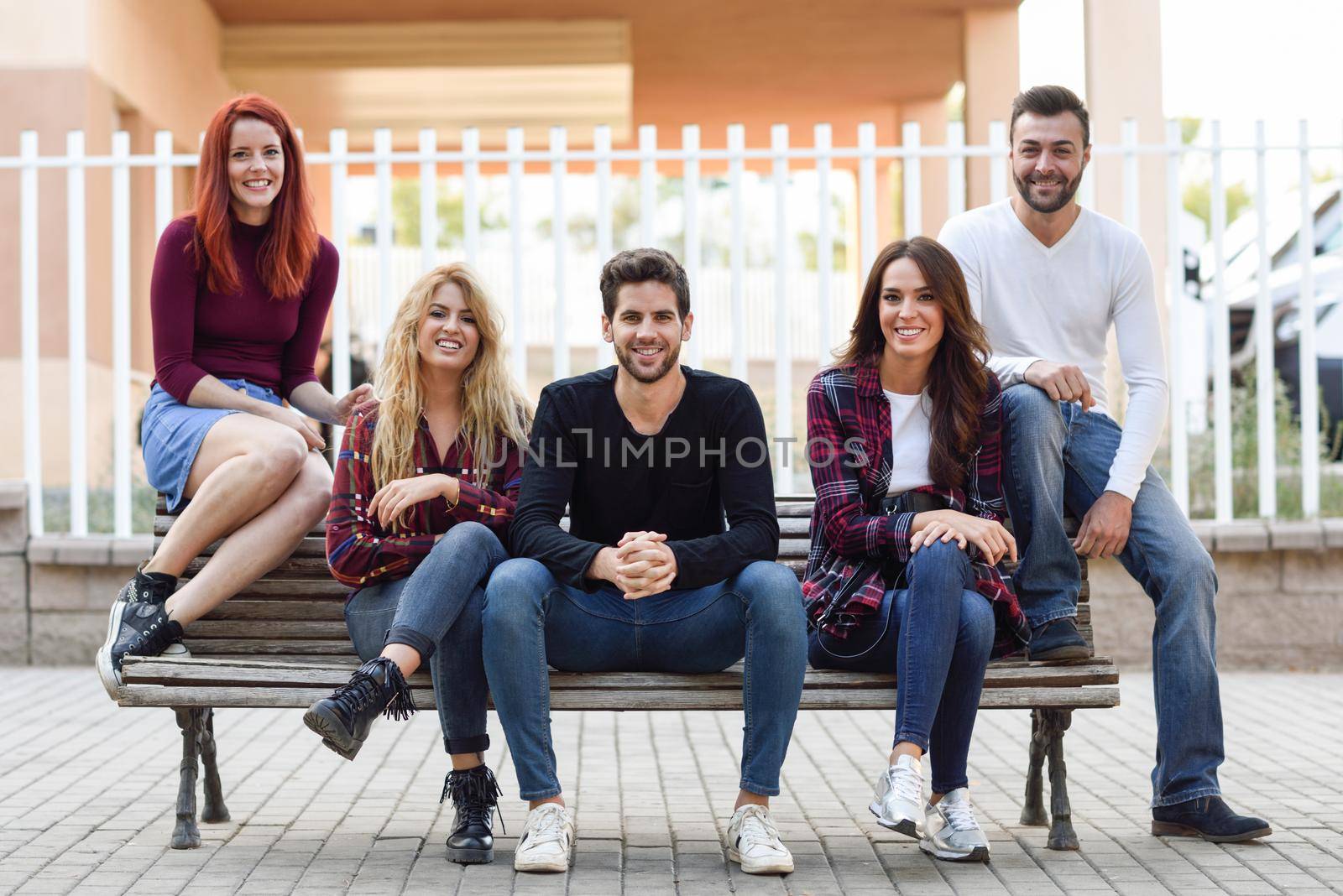 Group of young people together outdoors in urban background. Women and men sitting on a bench in the street wearing casual clothes.