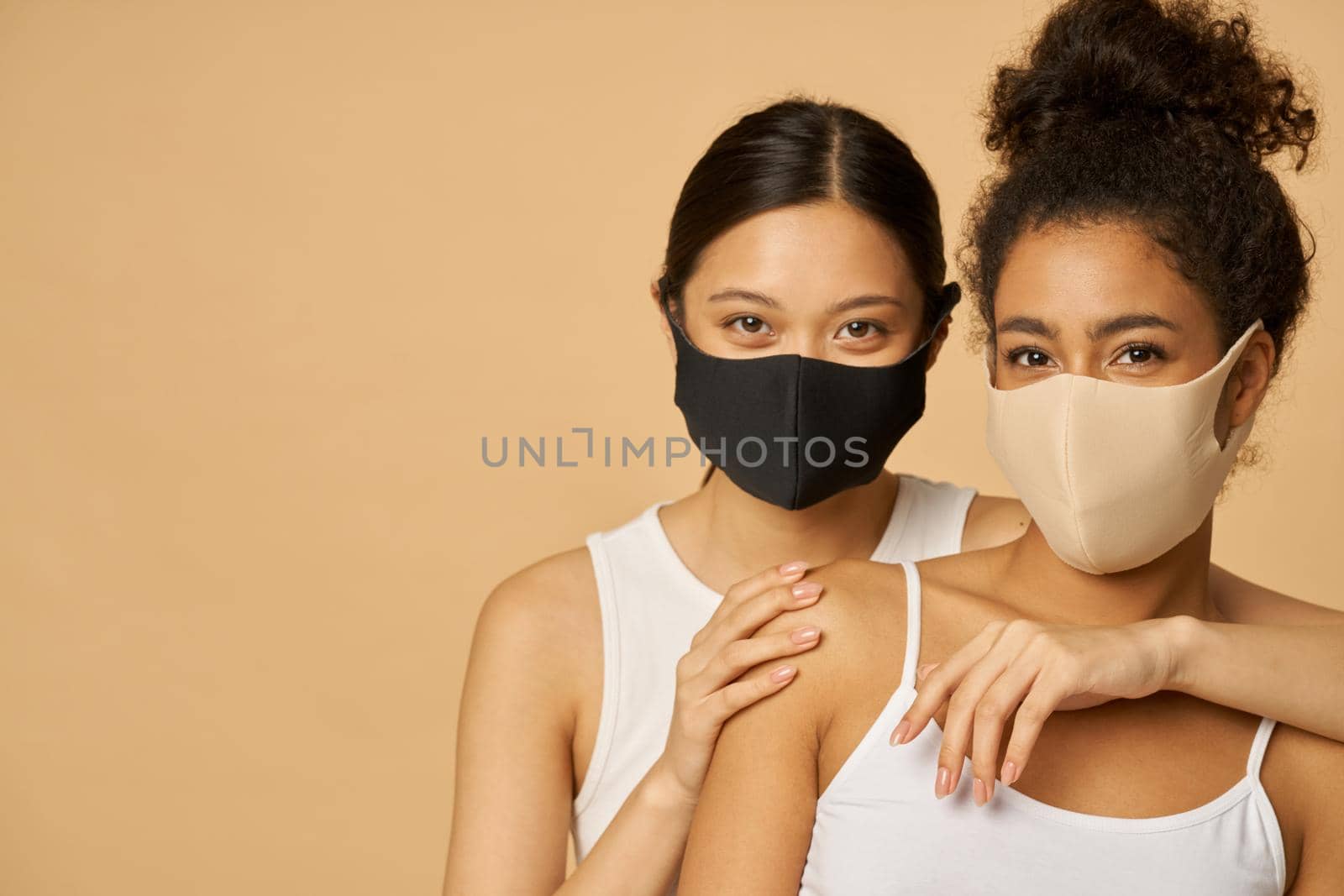 Portrait of two beautiful young diverse women wearing protective facial masks looking at camera while posing together isolated over beige background. Safety, pandemic concept