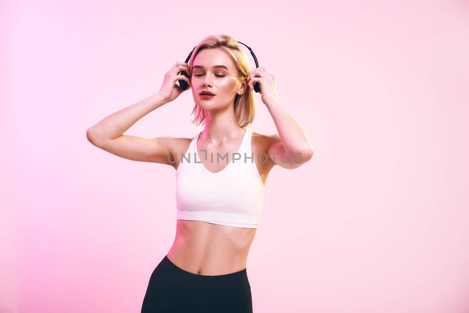 My favourite playlist. Cute and sporty woman adjusting headphones and keeping eyes closed while standing against pink background. Sport. Music concept